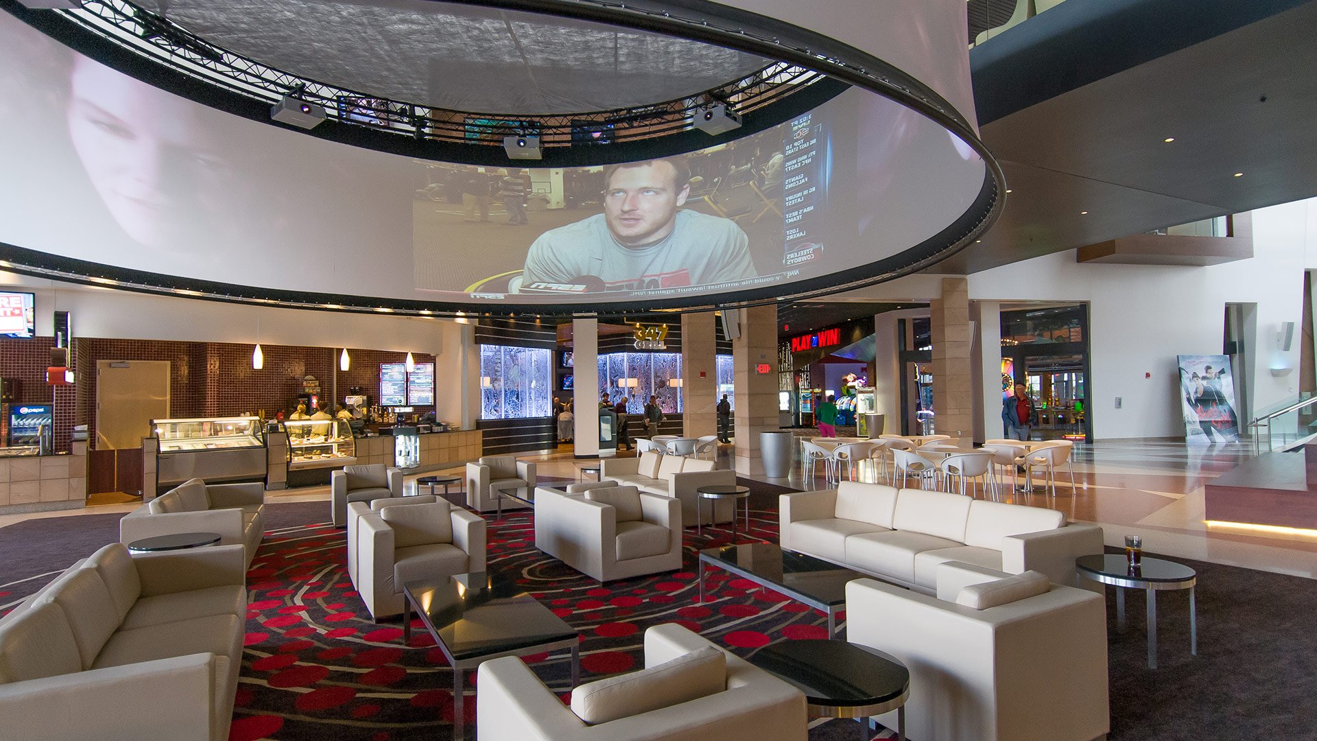 Interior of a movie theater lobby with seating area, food counters, and a large circular screen hanging from the ceiling displaying a man being interviewed.