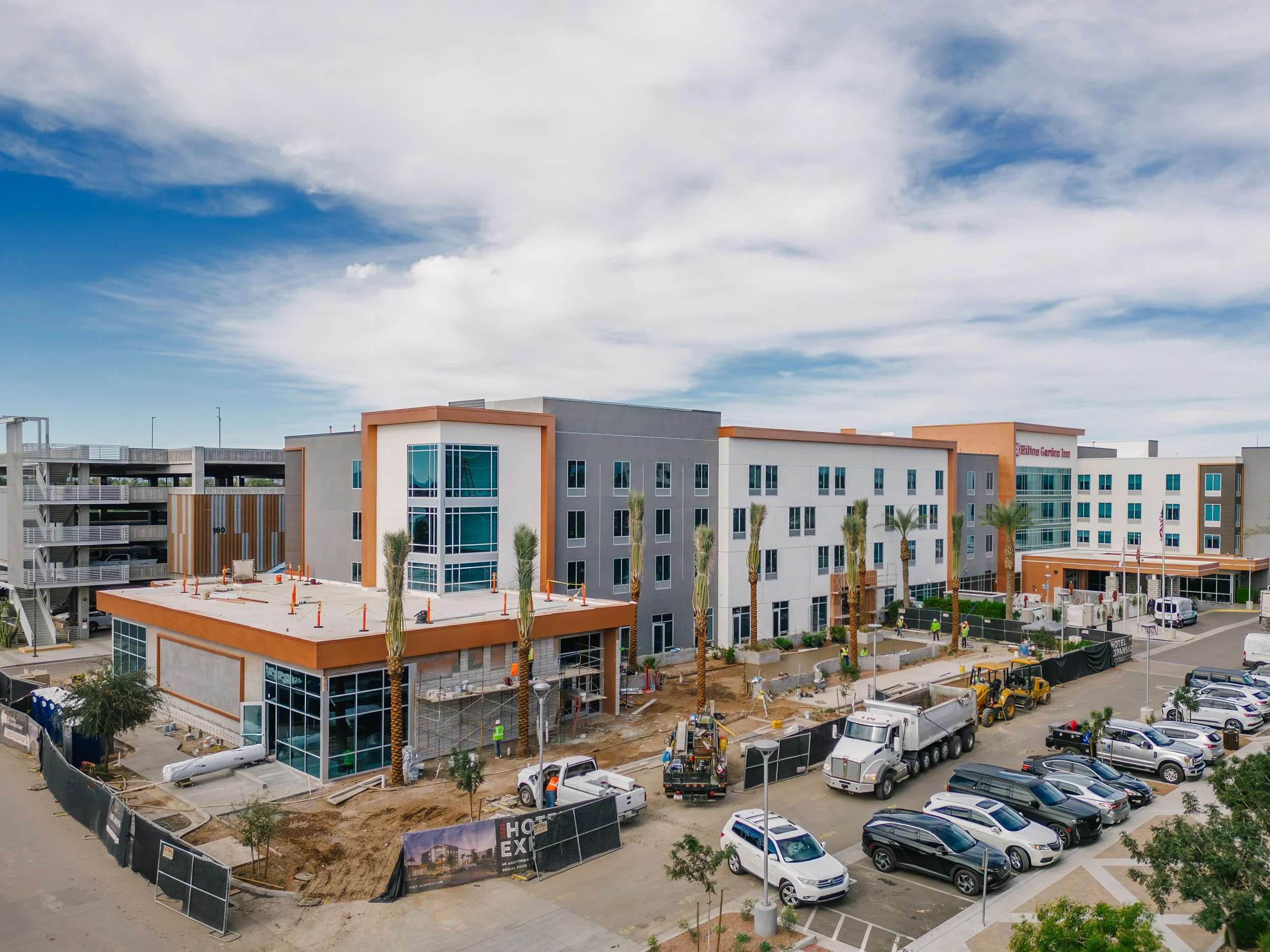 New multi-story building under construction with construction vehicles and parked cars in front, and a partly cloudy sky above.