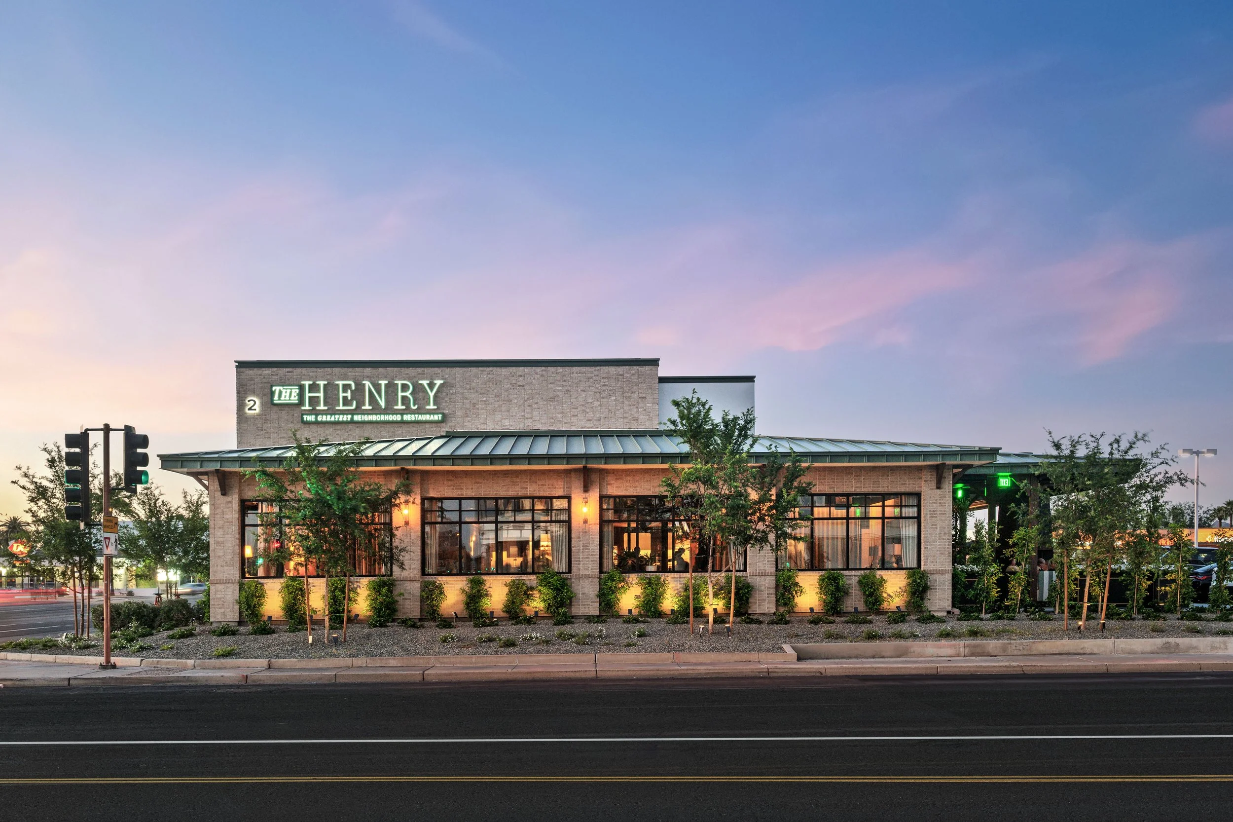 Exterior view of The Henry restaurant at dusk with trees and street in front, illuminated sign on building.