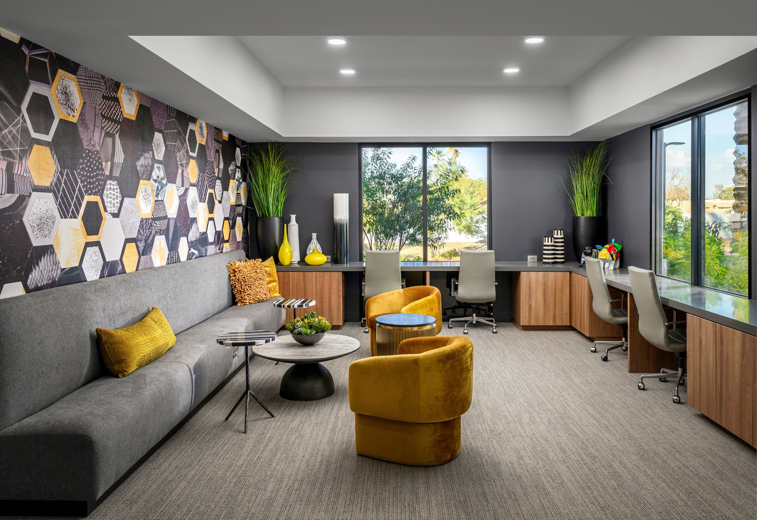 A modern office lounge with a gray sofa, yellow and textured throw pillows, two small black accent tables, yellow velvet chairs, a long windowed desk with three chairs, decorative vases, tall green plants, wallpaper with hexagon patterns, and a large