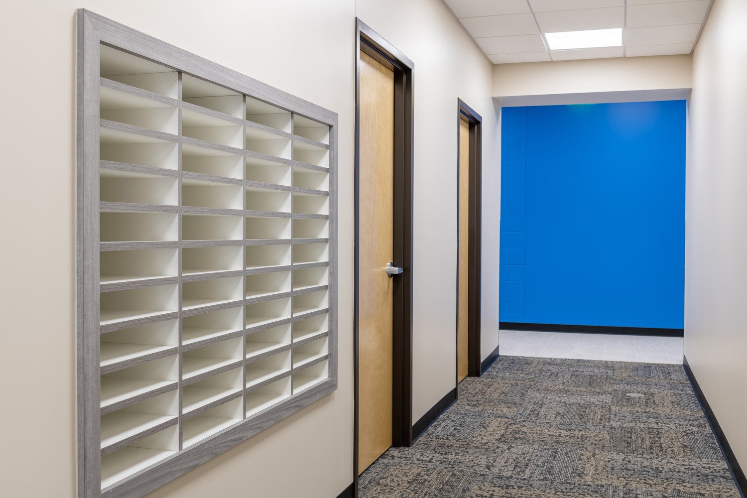 Empty mailroom with mail slots on wall, closed wooden doors, blue accent wall, and patterned carpet in hallway.