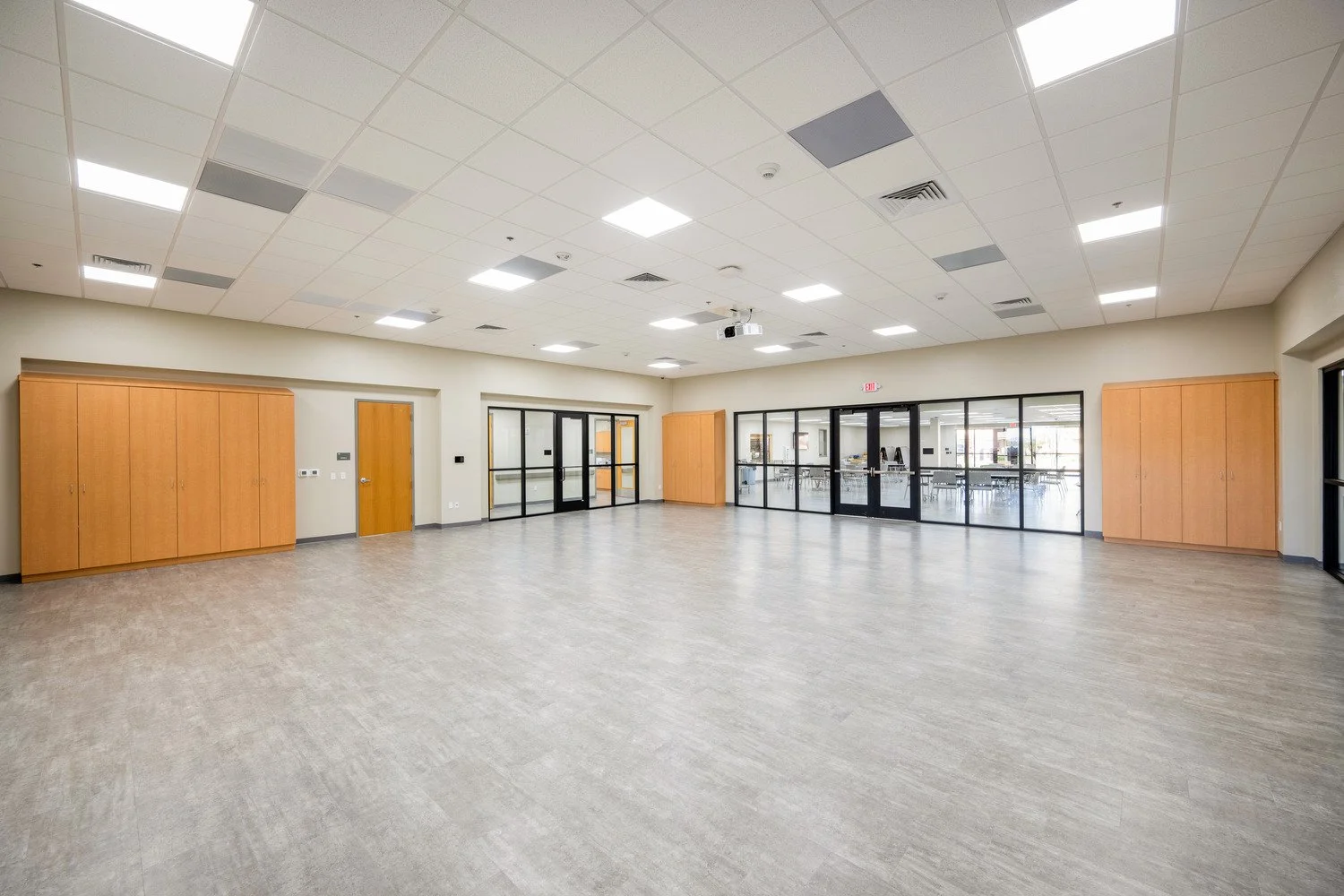 Empty multipurpose room with wooden cabinets, glass doors, drop ceiling with lights, and a view into a smaller room with tables and chairs.
