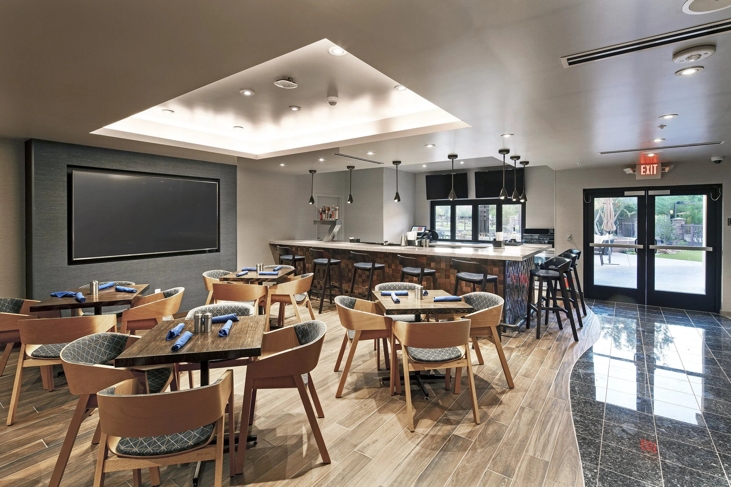 Upscale restaurant with wooden tables and chairs, a bar area with bar stools, a large TV screen on the wall, and large windows/doors leading outside.