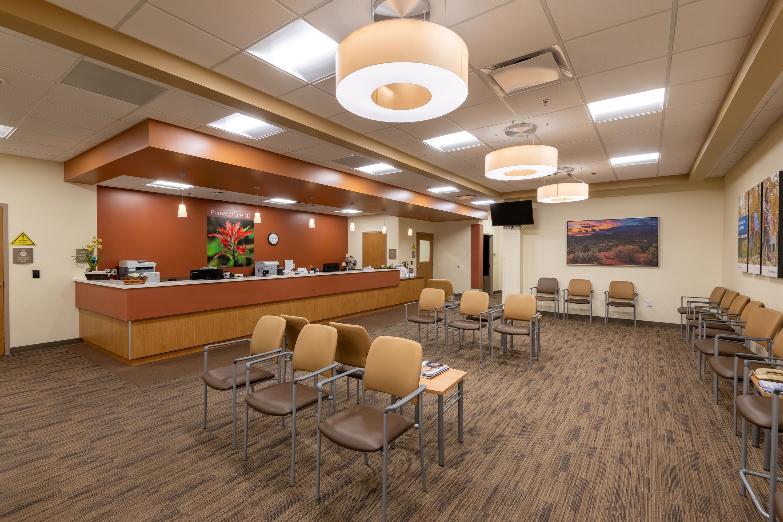 Empty hospital waiting room with rows of beige chairs, a reception desk with computers, and wall art.