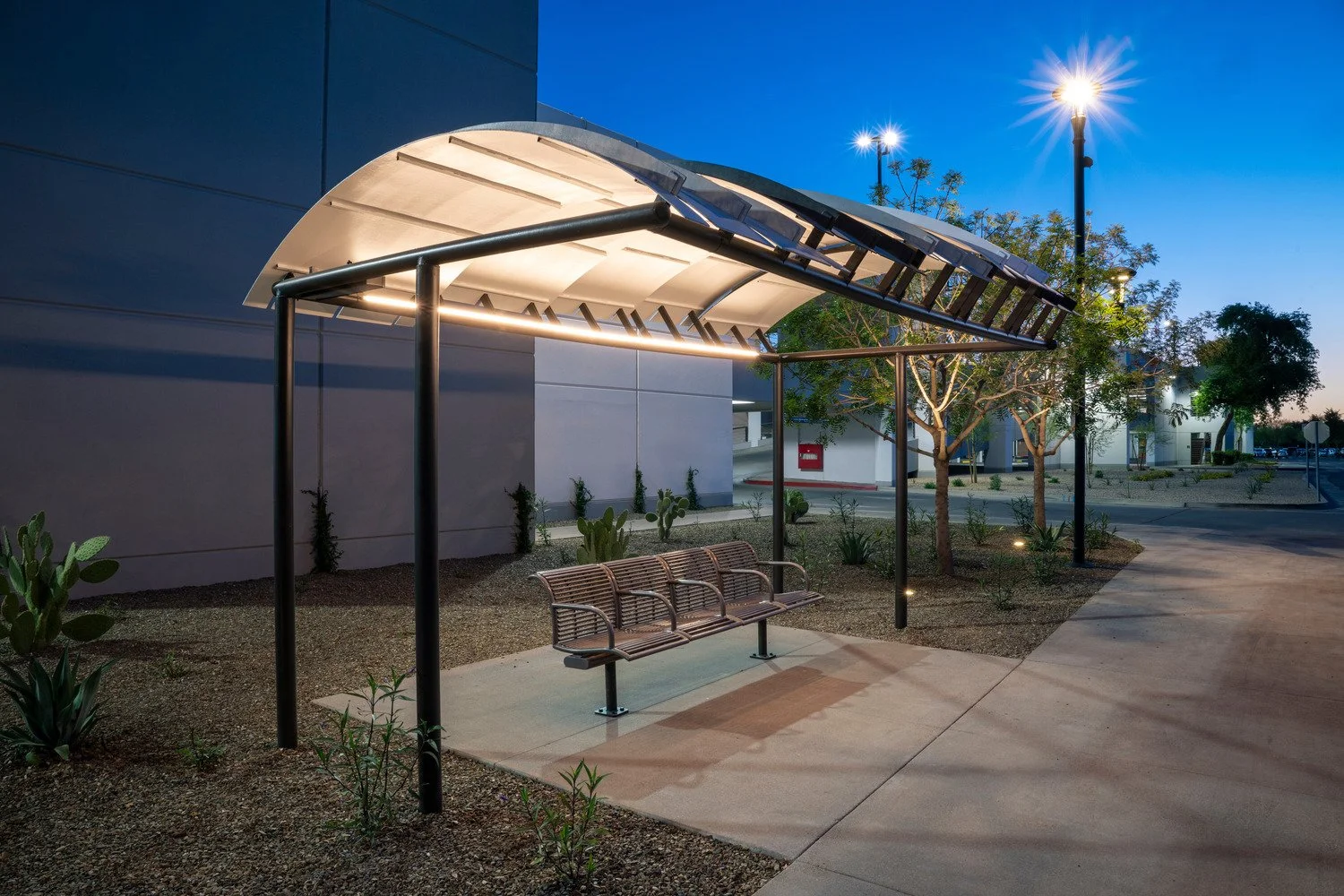 Empty outdoor bus shelter at dusk with wooden bench, illuminated roof, and surrounding trees and plants.