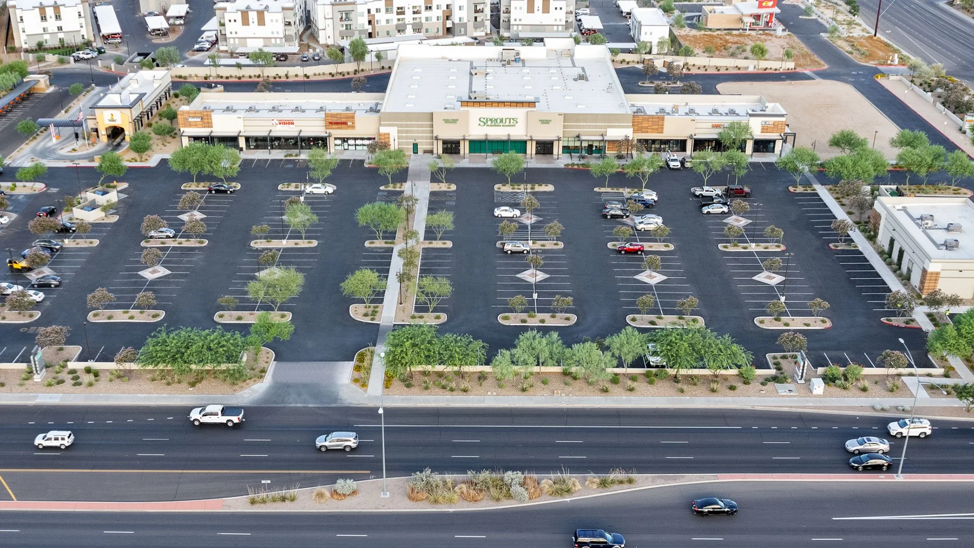 An aerial view of a shopping plaza with multiple storefronts, a large parking lot with some cars, landscaped areas with trees and bushes, and a busy street in front with moving vehicles.