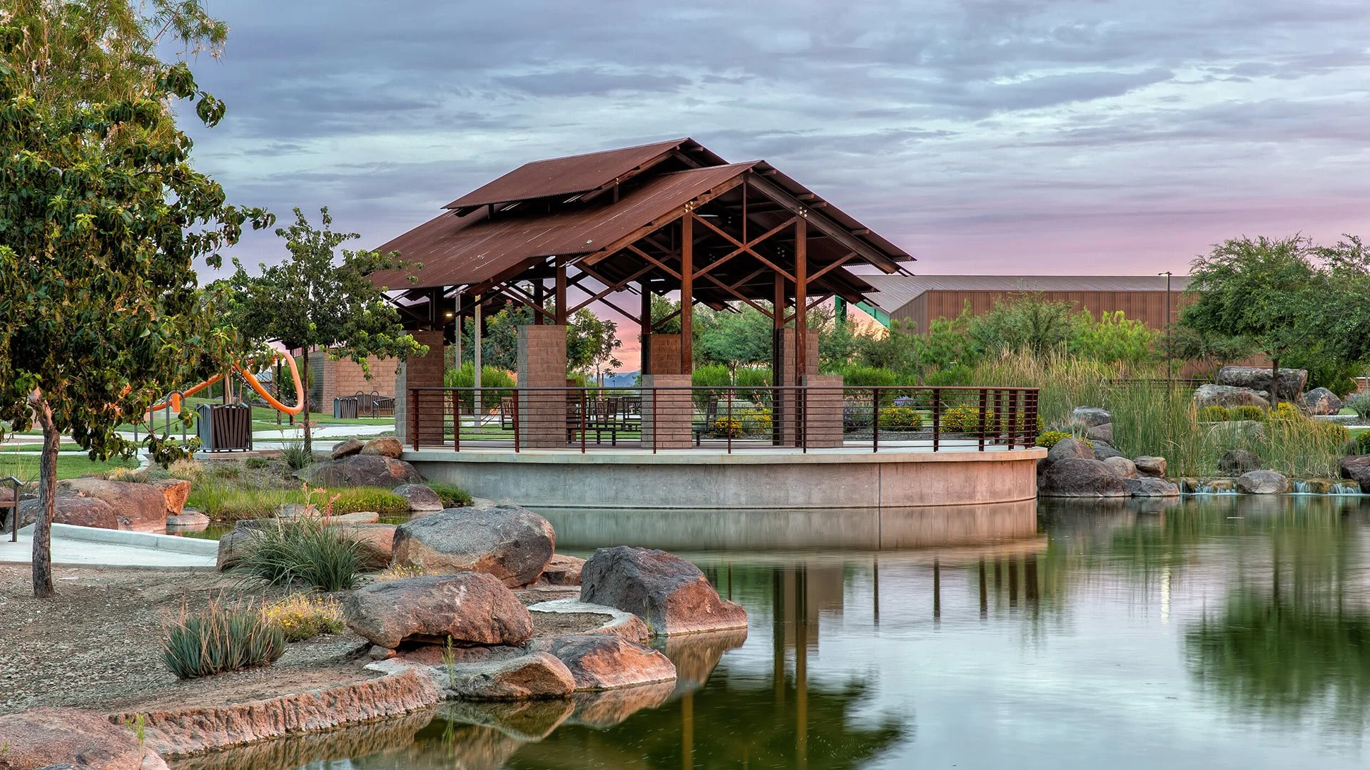 A park with a pond, trees, and rocks, featuring a wooden gazebo with a metal railing and a concrete base, during sunset with colorful sky.