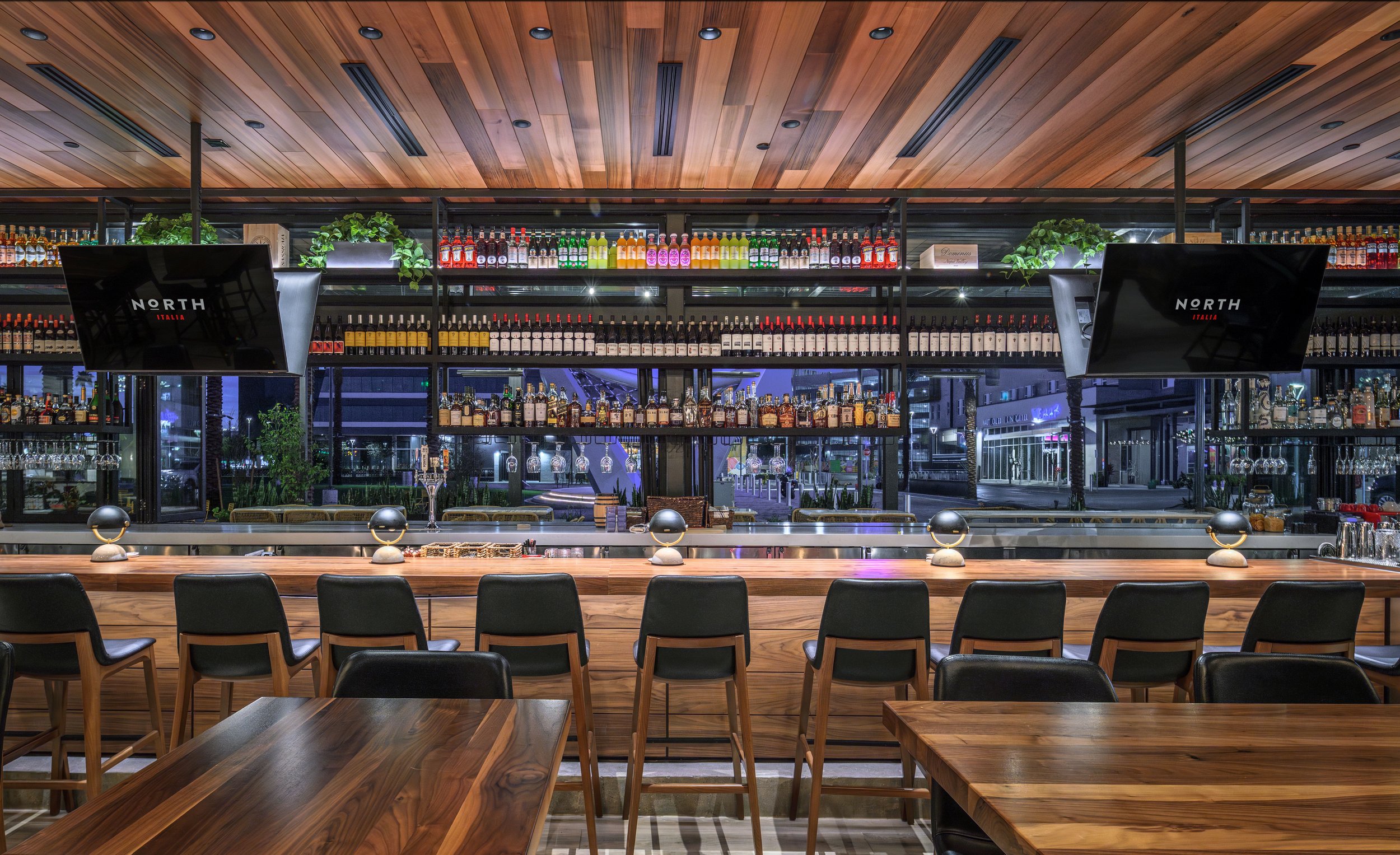 Modern indoor bar with a wooden counter, black chairs, and a back wall filled with shelves of various liquor bottles, illuminated by ceiling lights, with two mounted televisions displaying 'NORTH ITALIA.'