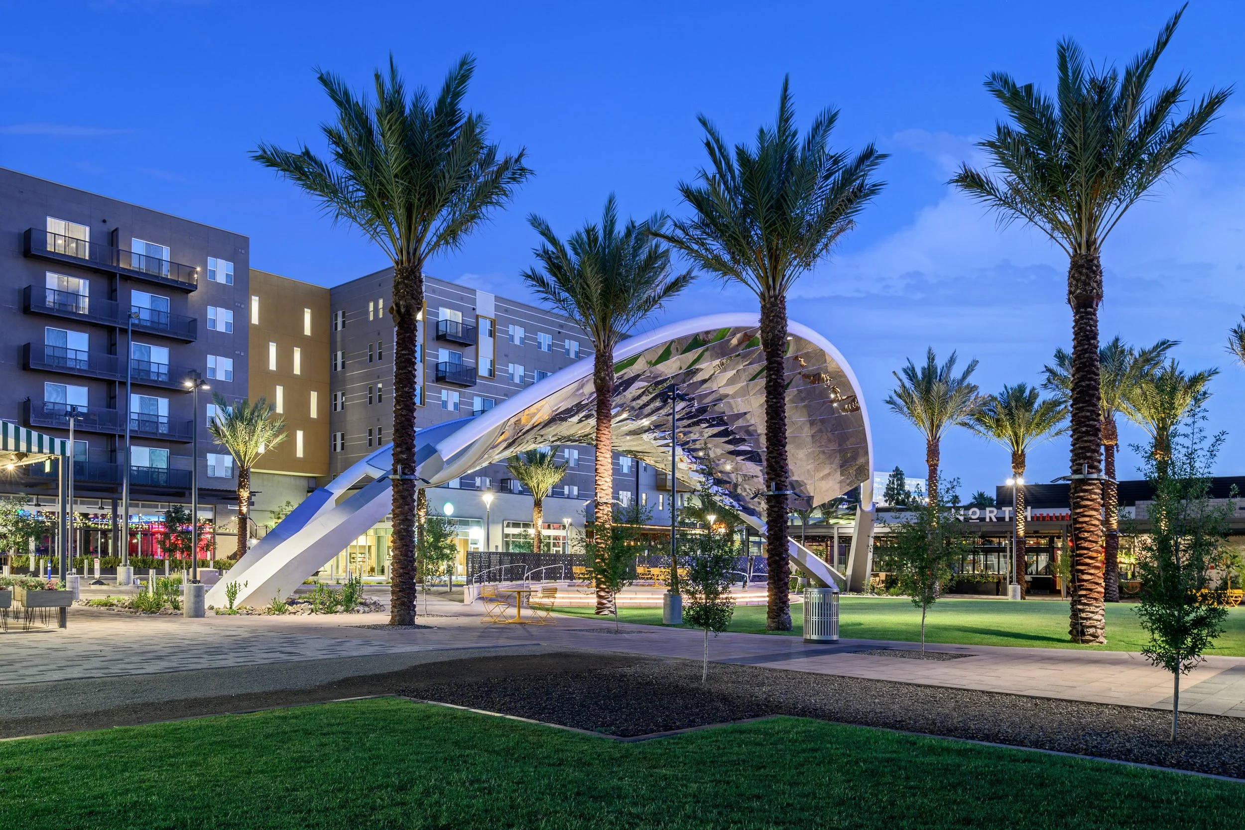 An outdoor public space at dusk with a modern architectural structure featuring a curved metallic roof, surrounded by tall palm trees and a multi-story building with lit windows in the background, illuminated by streetlights and landscaping.