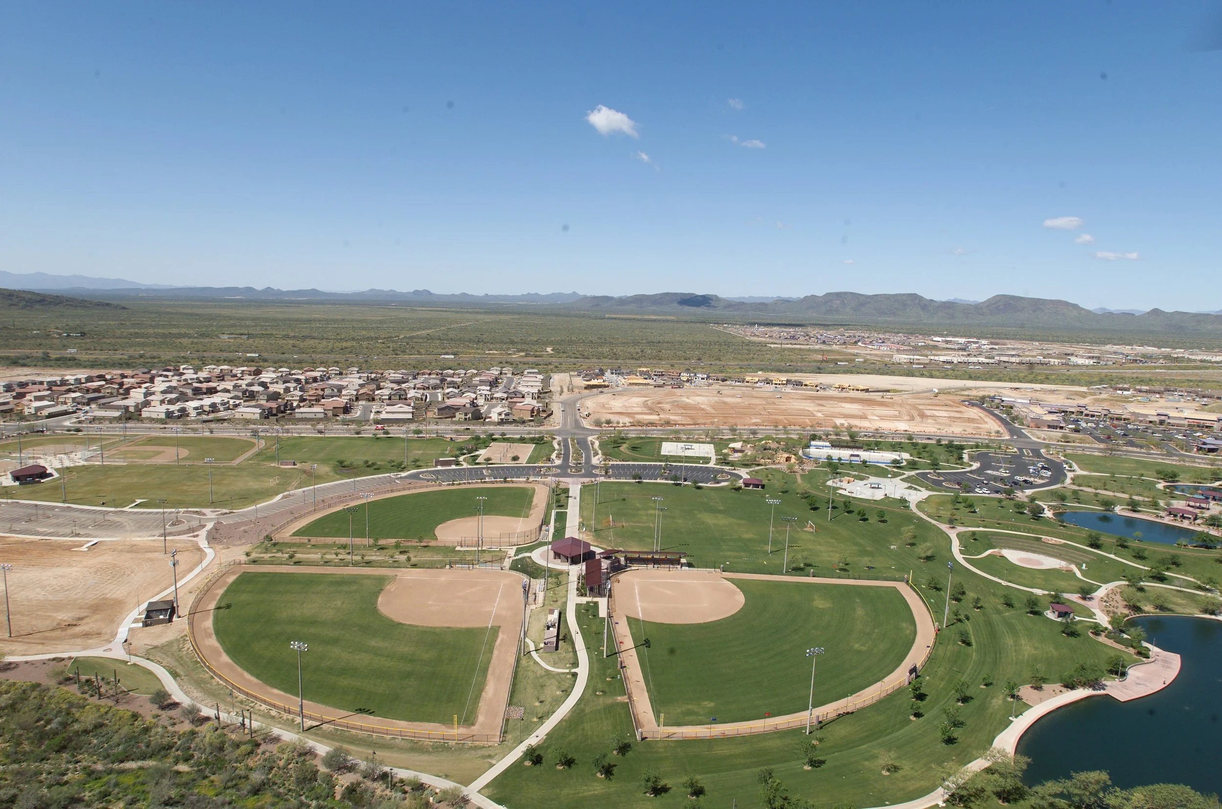 Aerial view of a park with baseball fields, walking paths, a pond, and a neighborhood in the background.