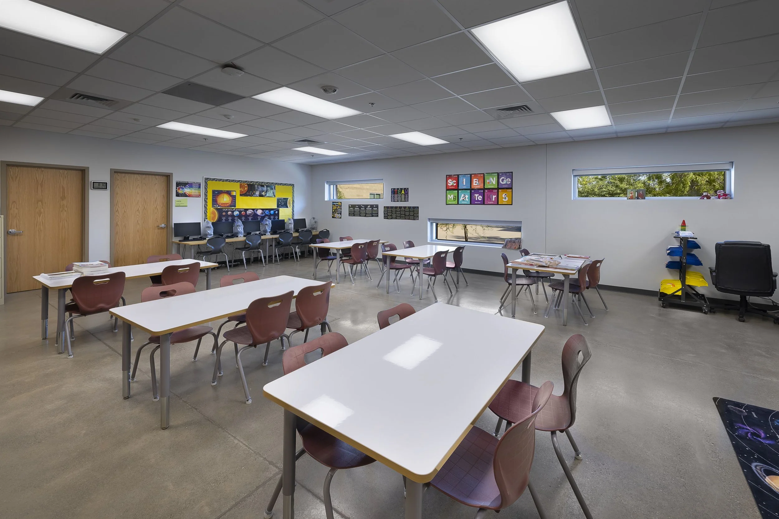 Empty classroom with white tables, brown chairs, windows, and educational posters on the walls.