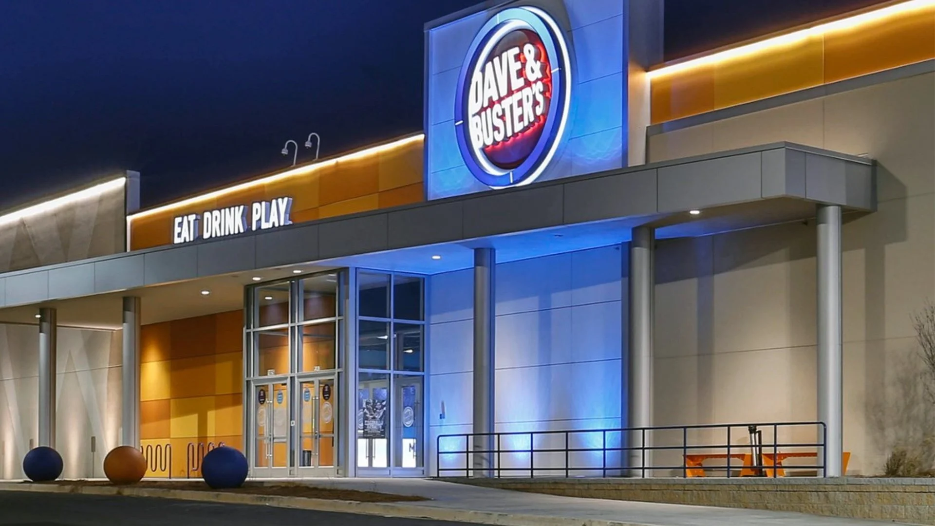 Night view of the exterior of a Dave & Buster's entertainment venue, with signage saying 'EAT DRINK PLAY', illuminated in blue and orange lights.