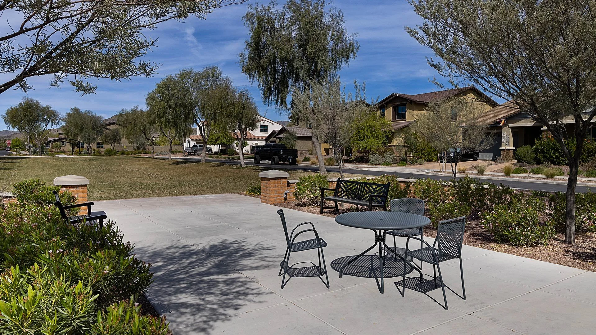 Park with metal table and chairs, benches, trees, residential houses, and blue sky.