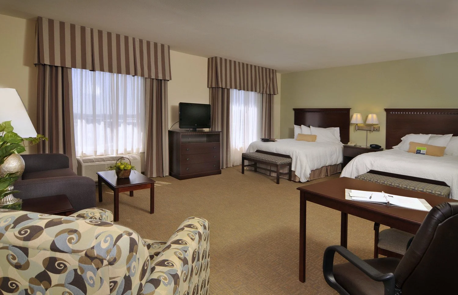 Hotel room with two beds, a tv on a dresser, seating area with patterned armchair and sofa, and a desk.