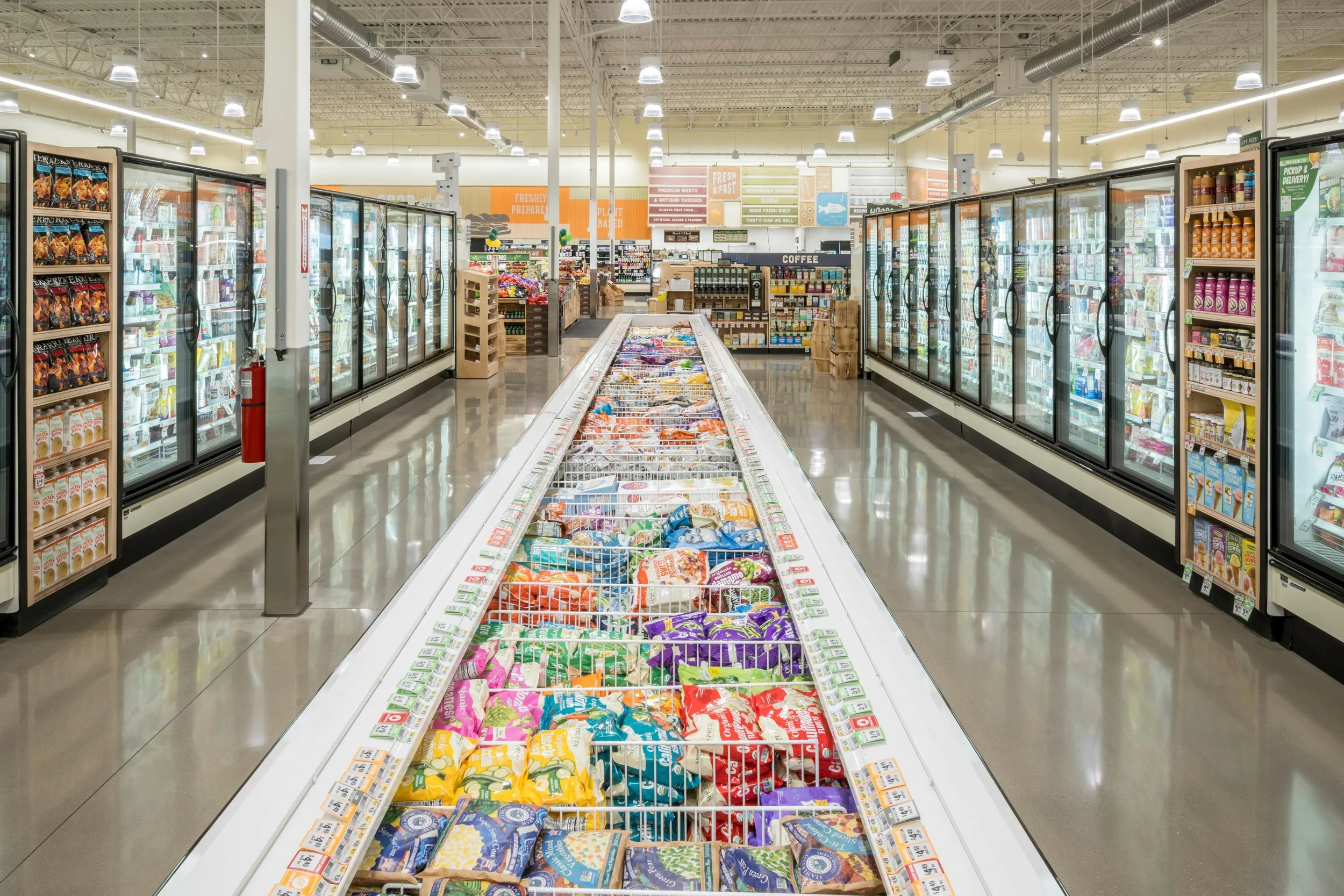 Inside a grocery store aisle with frozen foods in glass-door freezers on both sides, a low freezer with various packaged snacks in the center, and additional grocery sections in the background.
