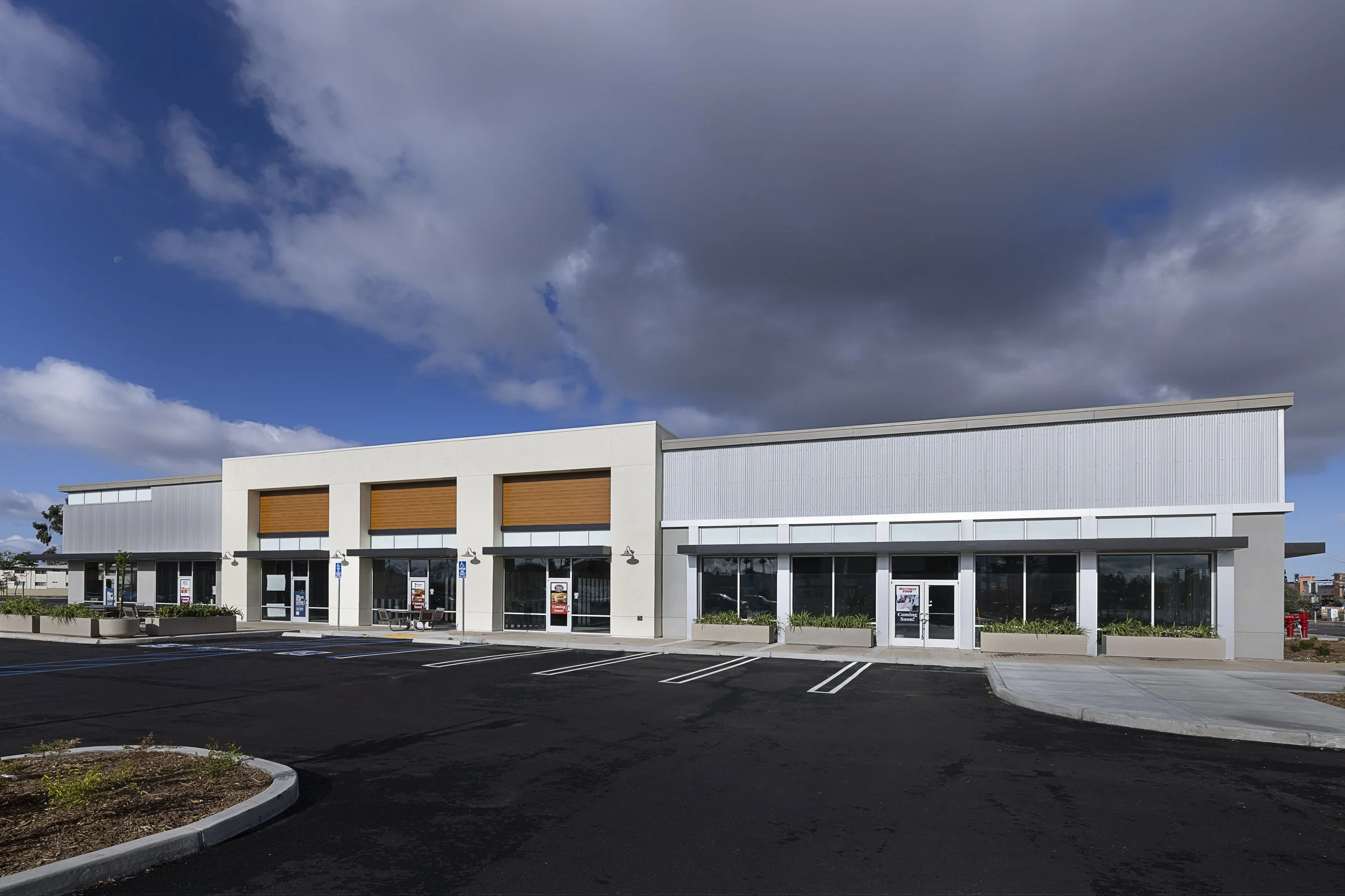 A modern commercial building with large glass windows and brown window shutters, situated in a parking lot with empty marked parking spaces. The sky is partly cloudy with a visible moon.