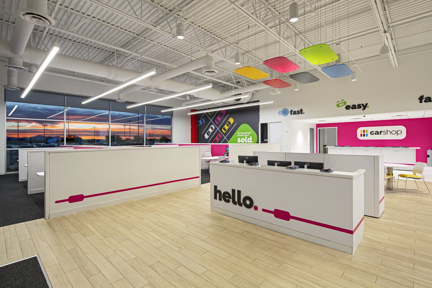 Interior of a modern car dealership with white desks, computer monitors, colorful ceiling panels, and promotional posters on the walls. Large windows show a sunset outside.