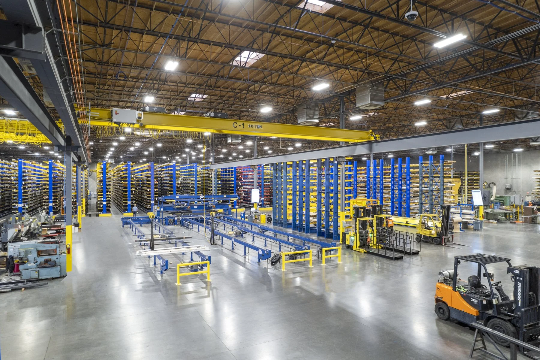 Interior of a large industrial warehouse with yellow overhead crane, blue and yellow shelving units, and machinery, including a forklift in the foreground.