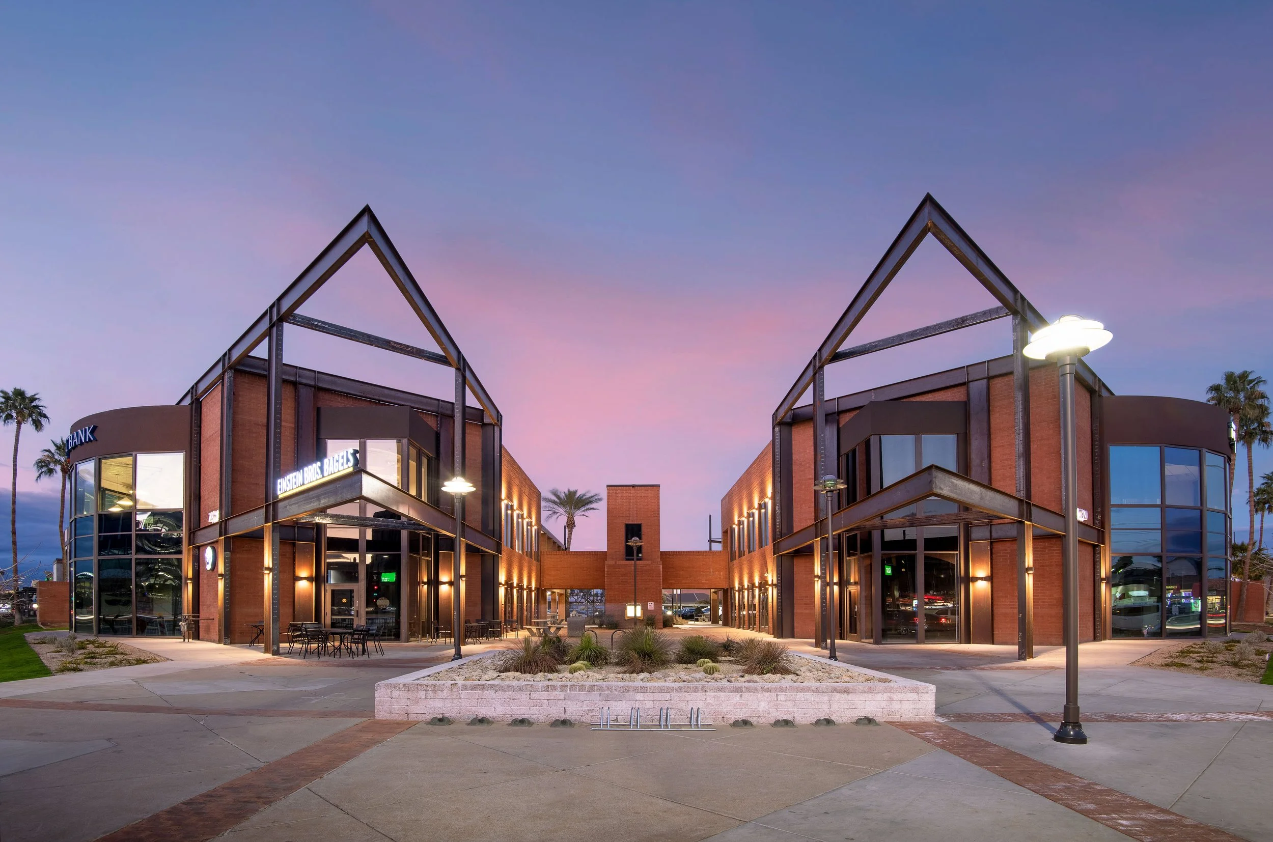 Two modern office buildings with large glass windows and exposed steel framing, illuminated at dusk, surrounded by palm trees and landscaped sidewalk.