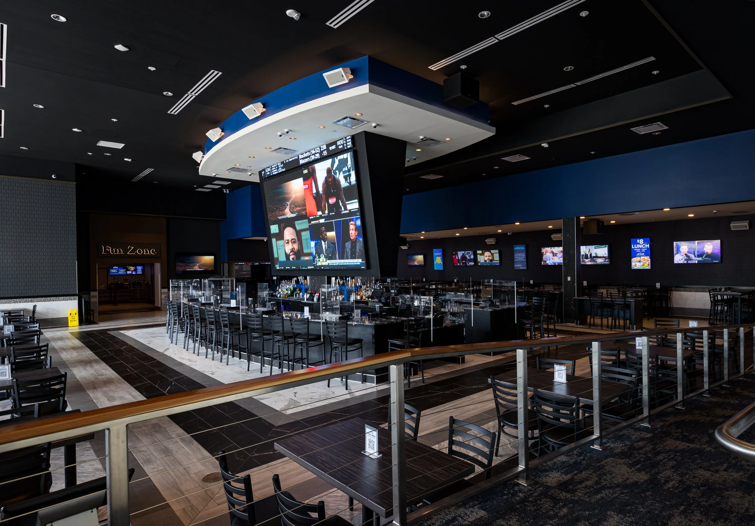 An empty sports bar with a large screen displaying multiple video feeds, a bar counter with stools, and several smaller screens on the walls, with no people present.