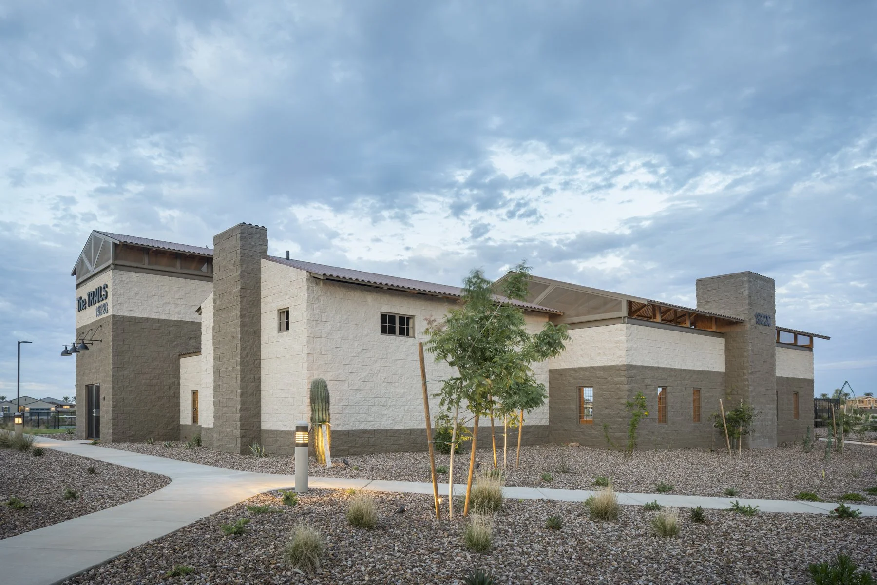 Modern residential building with beige and brown exterior walls, cacti, and desert landscaping, under a cloudy sky.