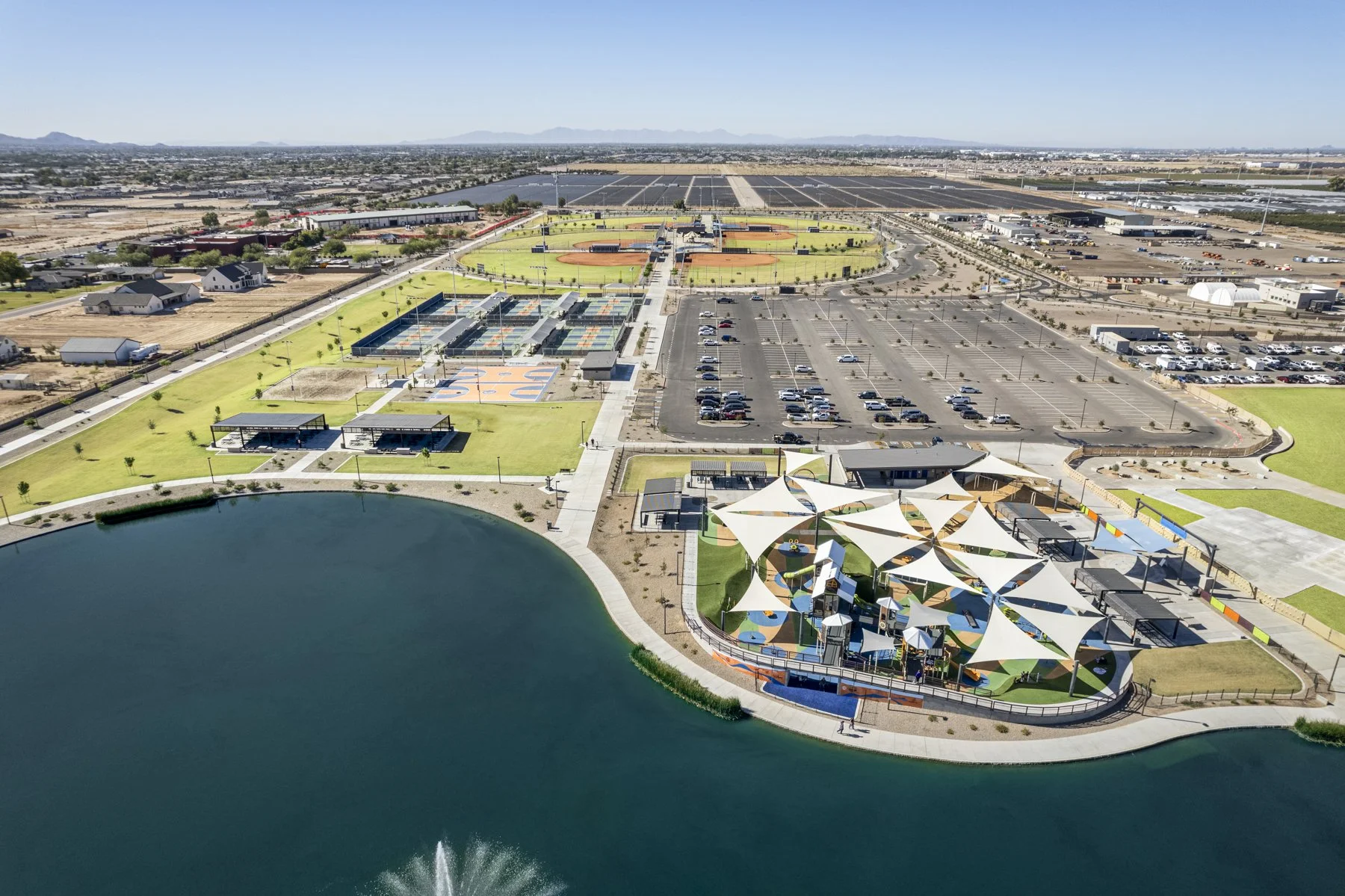 Aerial view of a large recreational park with a lake, playground area with shade sails, sports courts, and parking lot, surrounded by open land and distant mountains under a clear blue sky.