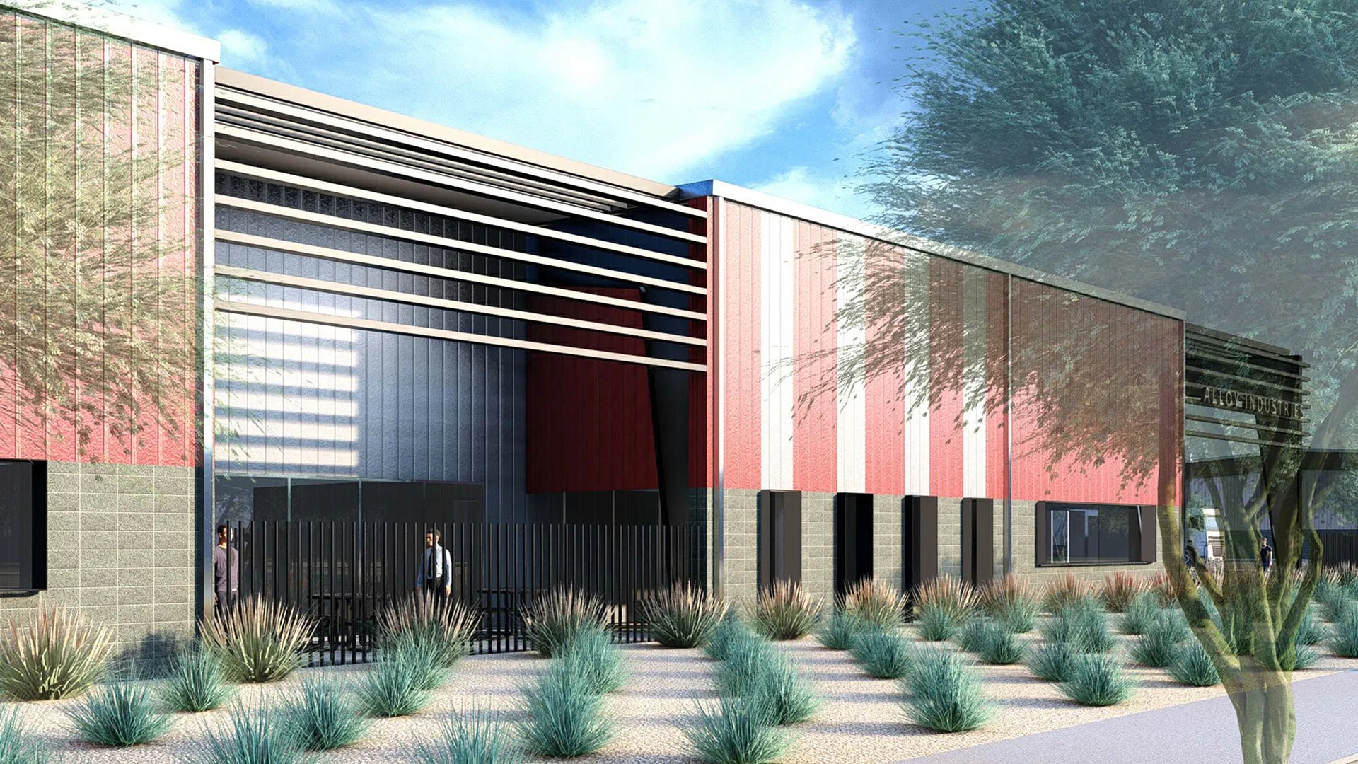 A modern industrial building with red and gray exterior, metal slatted overhang, and a black fence at the entrance. Landscaping includes desert plants and a tree, with a clear blue sky in the background.