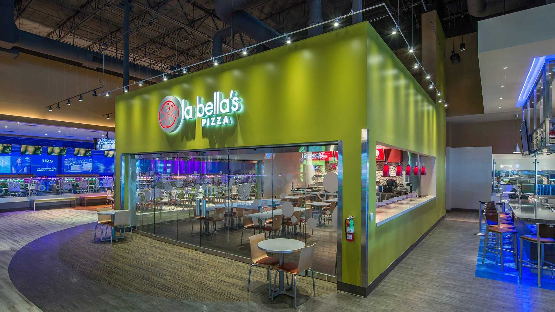 Interior of La Bella's Pizza restaurant in a mall or shopping center, with a green exterior wall, glass windows, seating area inside, red pendant lights, and a same-side counter with bar-style chairs.