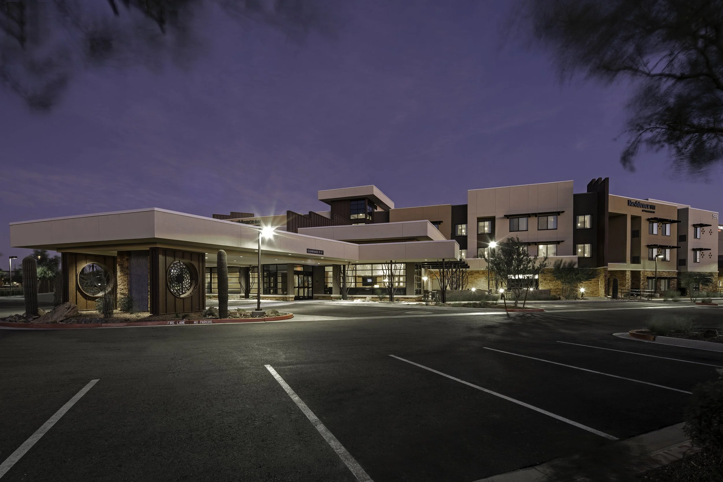 Nighttime exterior view of a modern hotel with a well-lit parking lot and a desert landscape featuring cacti and rocks.