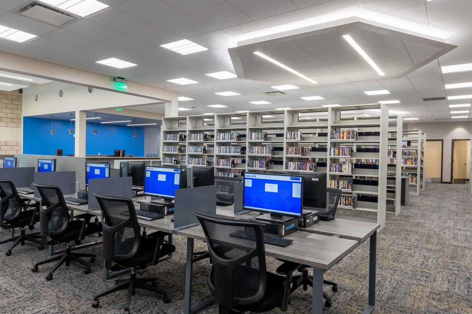 A computer lab with multiple desktop computers arranged on long tables, black ergonomic chairs, and bookshelves full of books. The ceiling has bright lights and there's a section with a blue wall and pendant lights in the background.