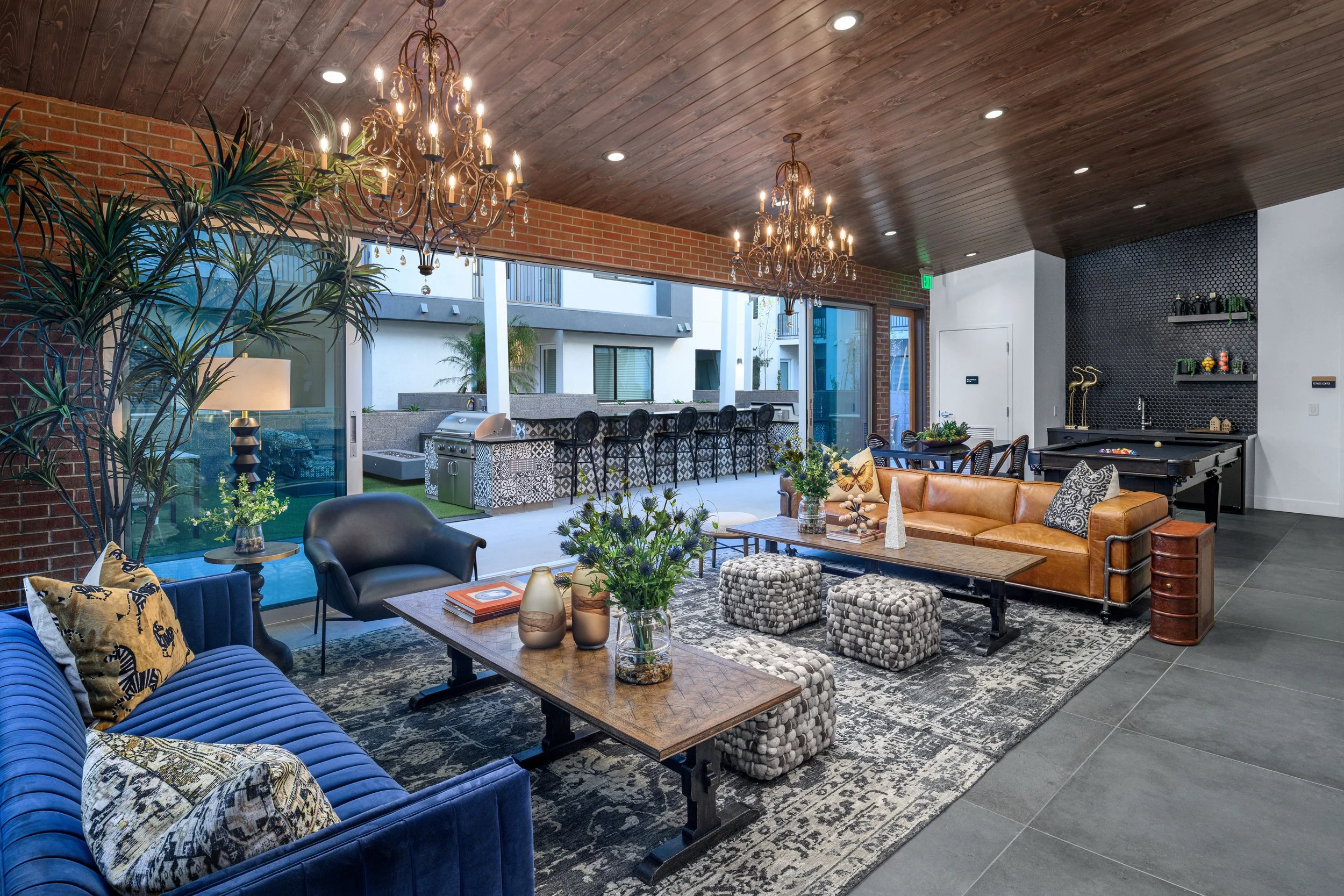 Living room with leather sofa, patterned ottomans, black armchair, wooden table with vases, large potted plant, chandelier, open kitchen with outdoor bar area, and sliding glass doors leading to patio.