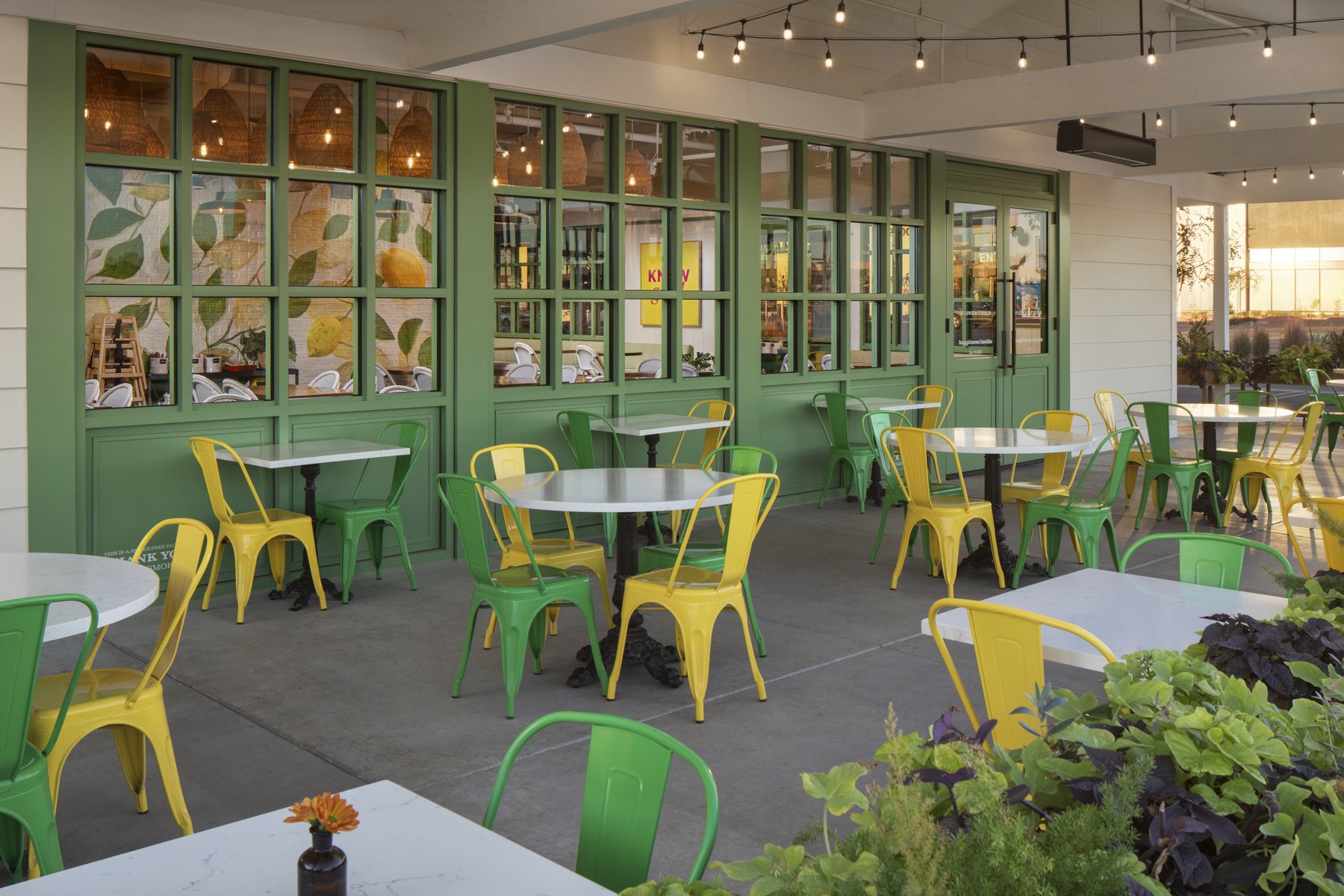 Empty outdoor cafe patio with white tables, green and yellow metal chairs, and a backdrop of a restaurant with green window frames and string lights.