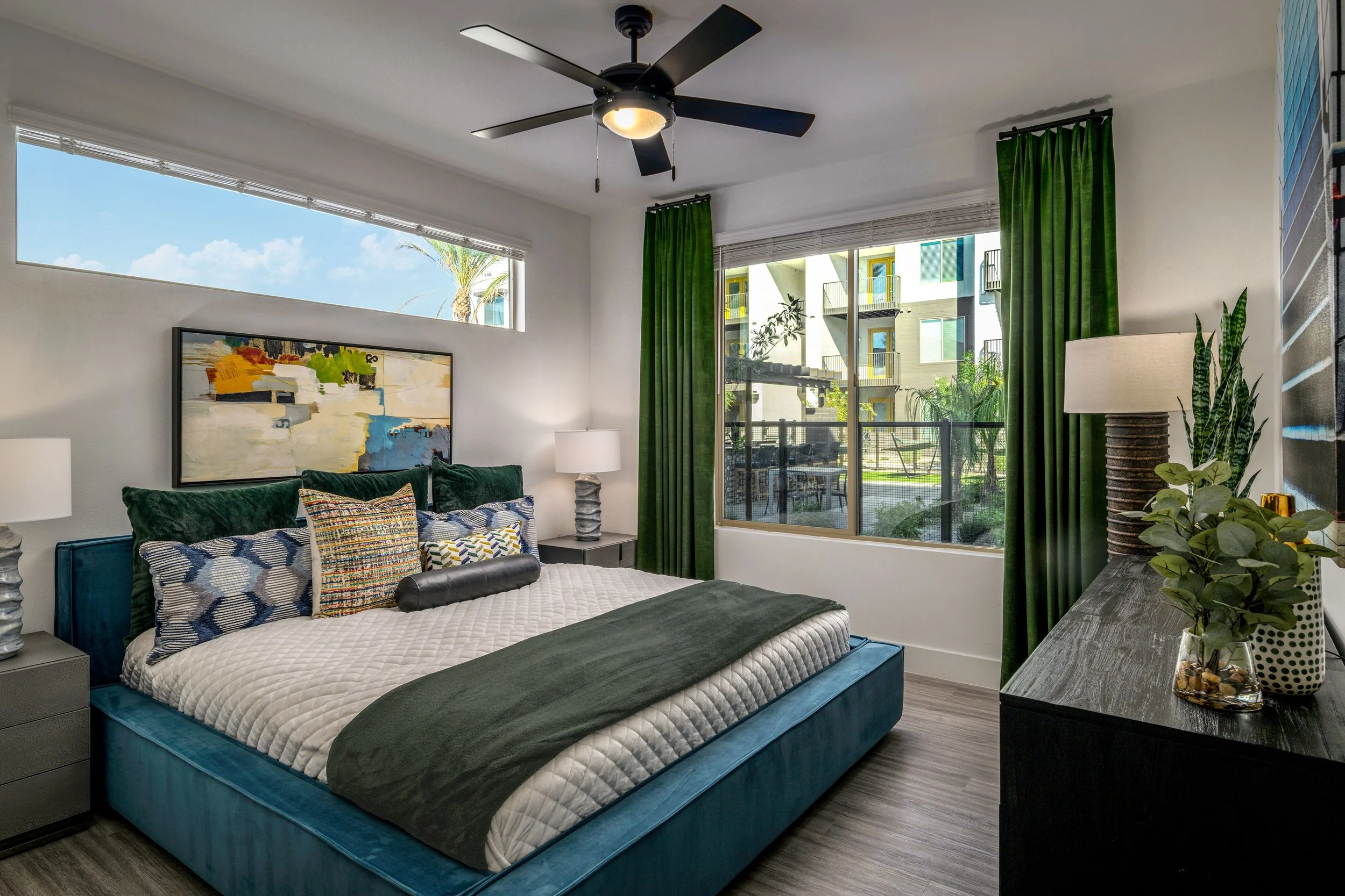 Modern bedroom with a large teal bed, green velvet pillows, and a white quilted blanket. There are two nightstands with lamps on either side of the bed, a large window with green curtains, and a balcony with outdoor furniture. A ceiling fan is above,