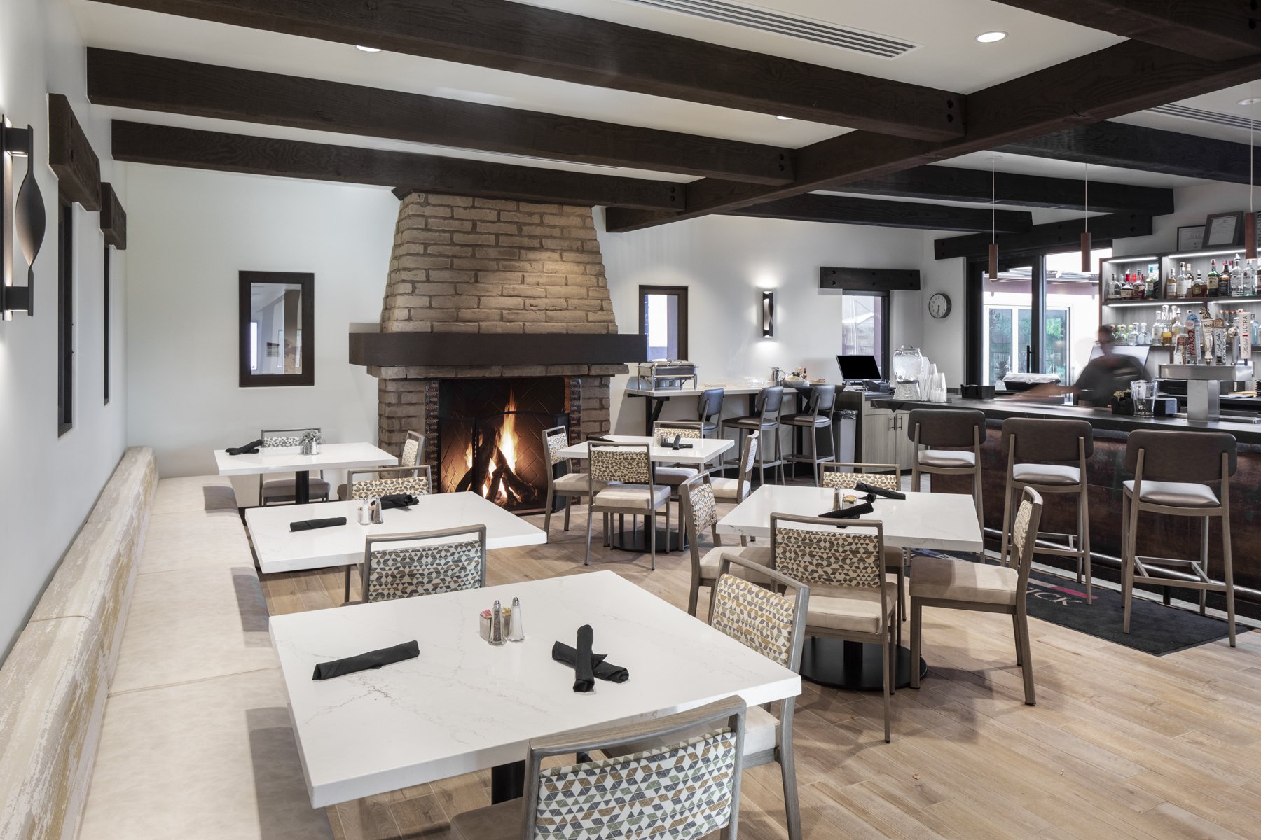 Cozy restaurant interior with a fireplace, white tables with black napkins, and bar area in the background.