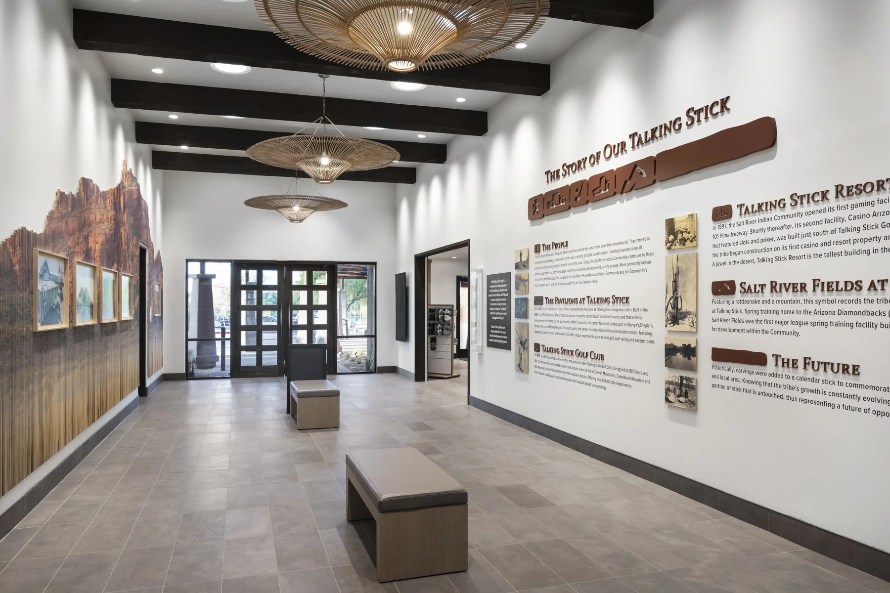 Modern inside lobby with white walls, wooden benches, wall plaques, and large artwork on the left wall, with a glass door and window at the back and decorative ceiling lights.