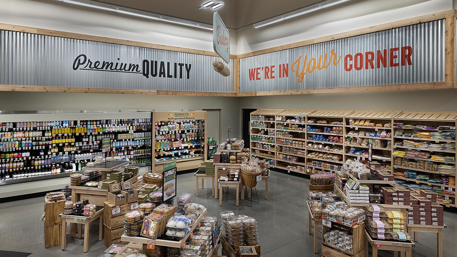 Interior of a grocery store aisle with shelves stocked with baked goods, snacks, and pantry items, and a sign overhead reading 'Premium Quality' and 'We're in Your Corner'.