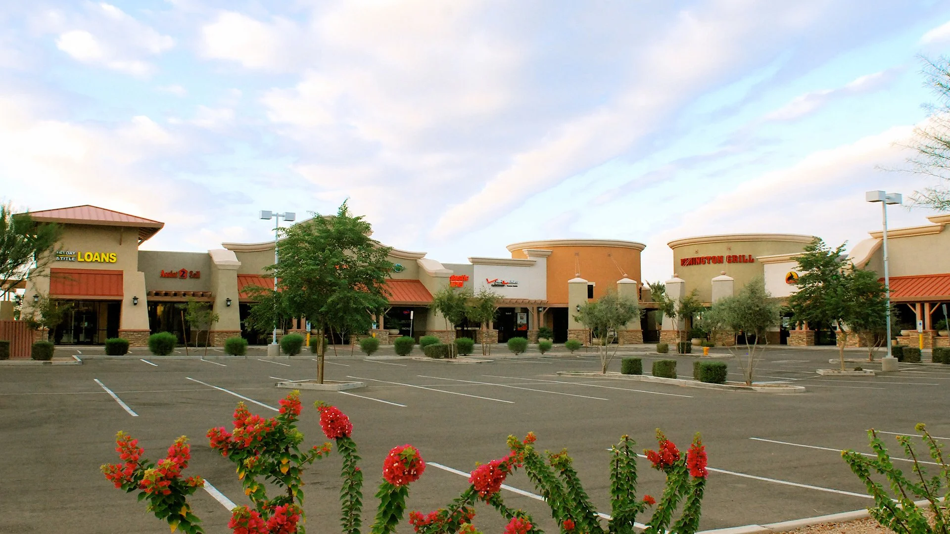 Empty parking lot in front of a shopping plaza with trees and flowers, under a partly cloudy sky.