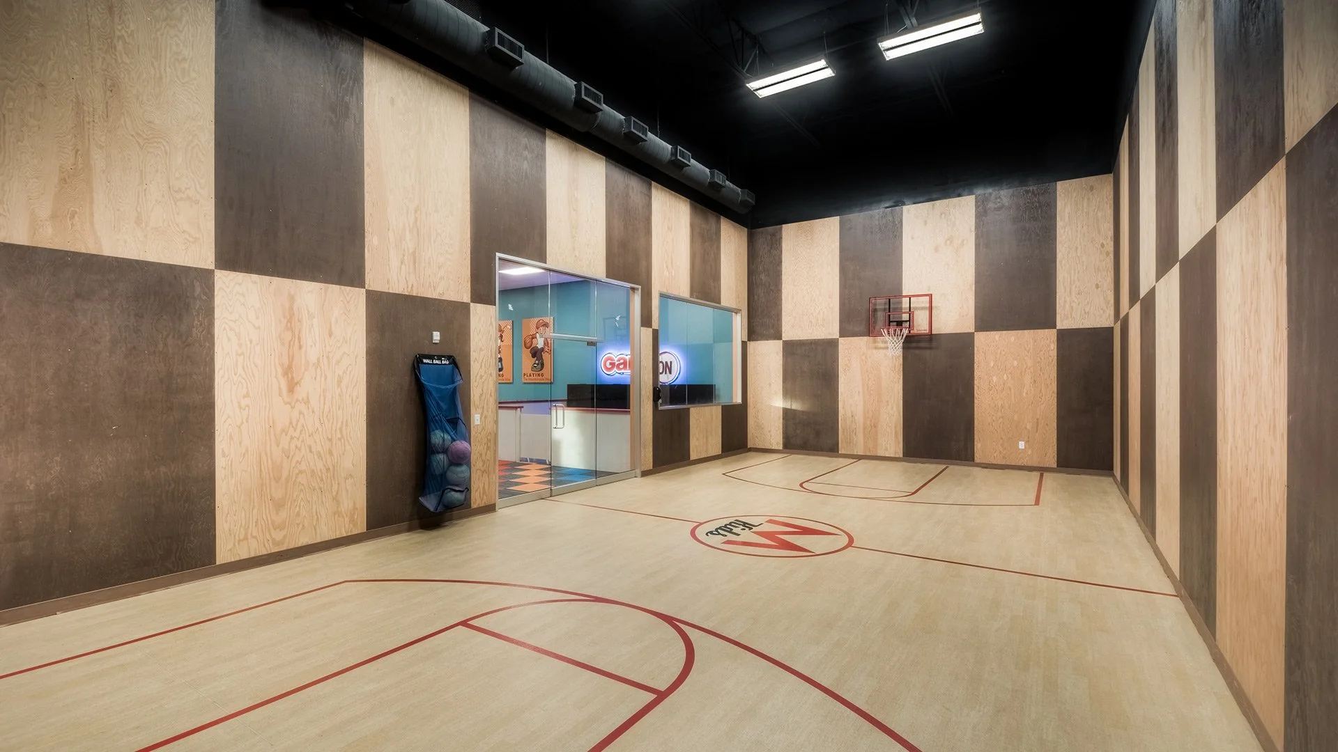 Empty indoor basketball gym with wood panel walls, basketball hoop, and a wall-mounted ball storage net.