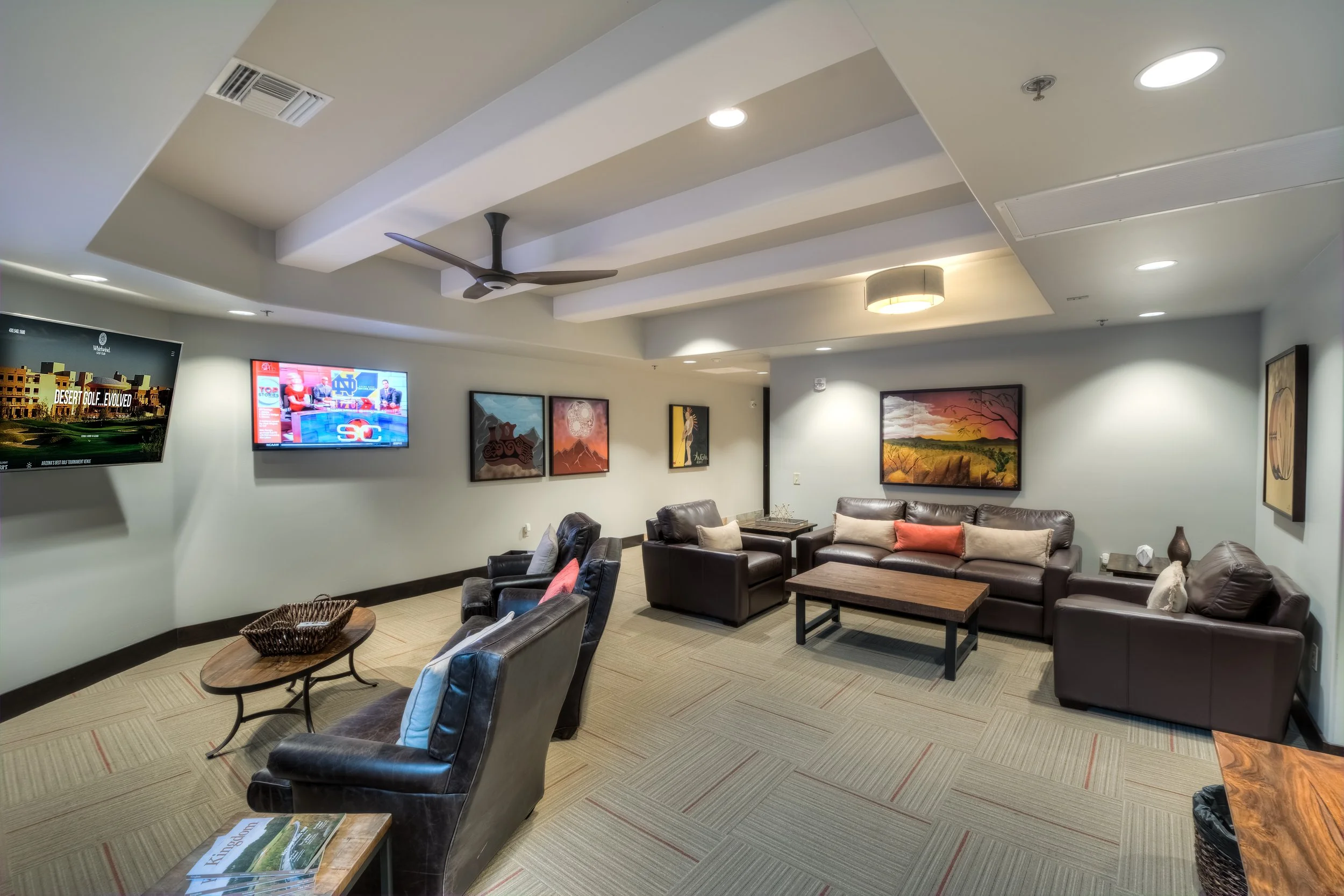 Living room with beige carpet, dark leather sofas and chairs, wall-mounted TVs, colorful paintings, ceiling fan, and modern lighting.