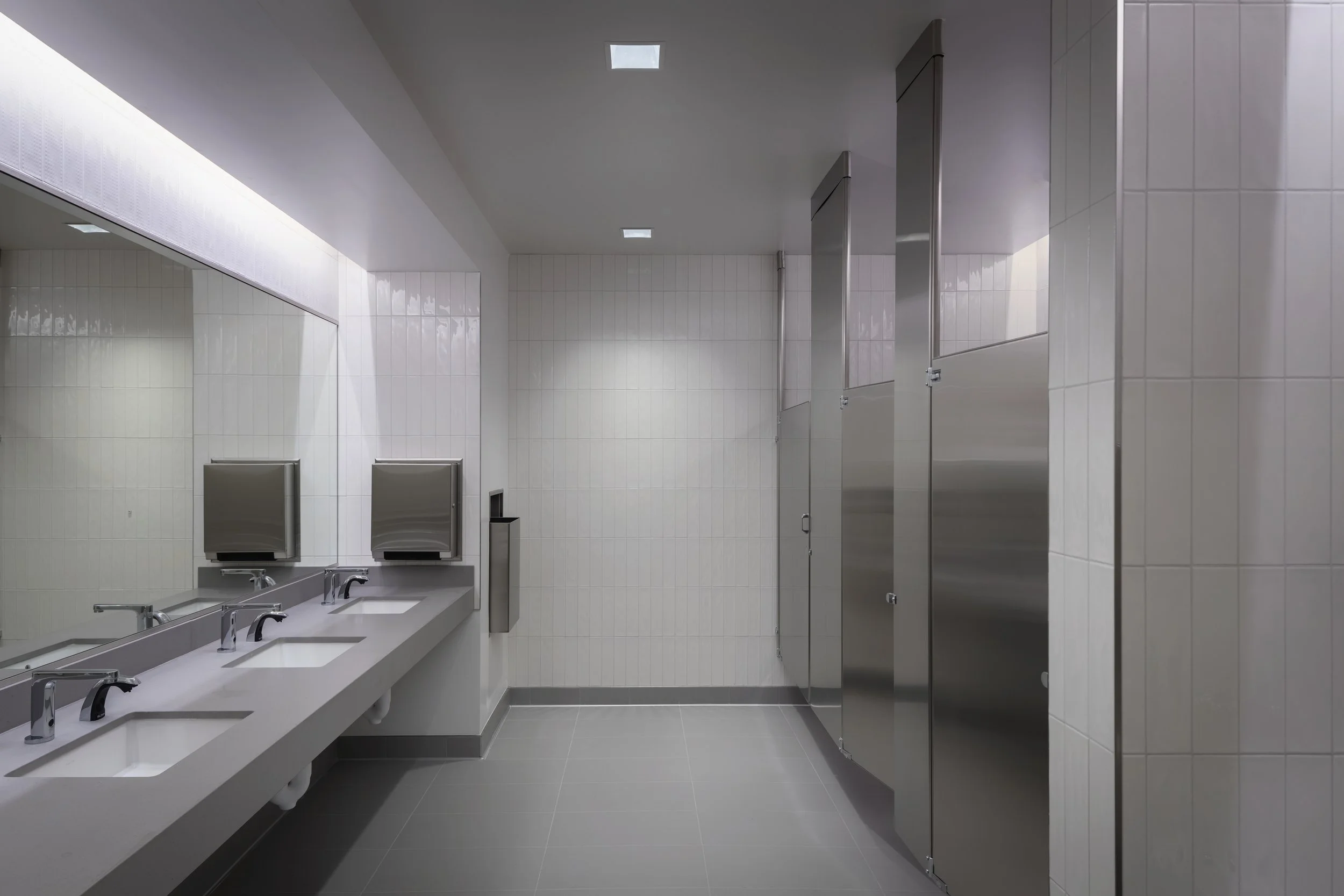 Public restroom with three sinks, a large mirror, soap dispensers, and stalls with stainless steel doors.