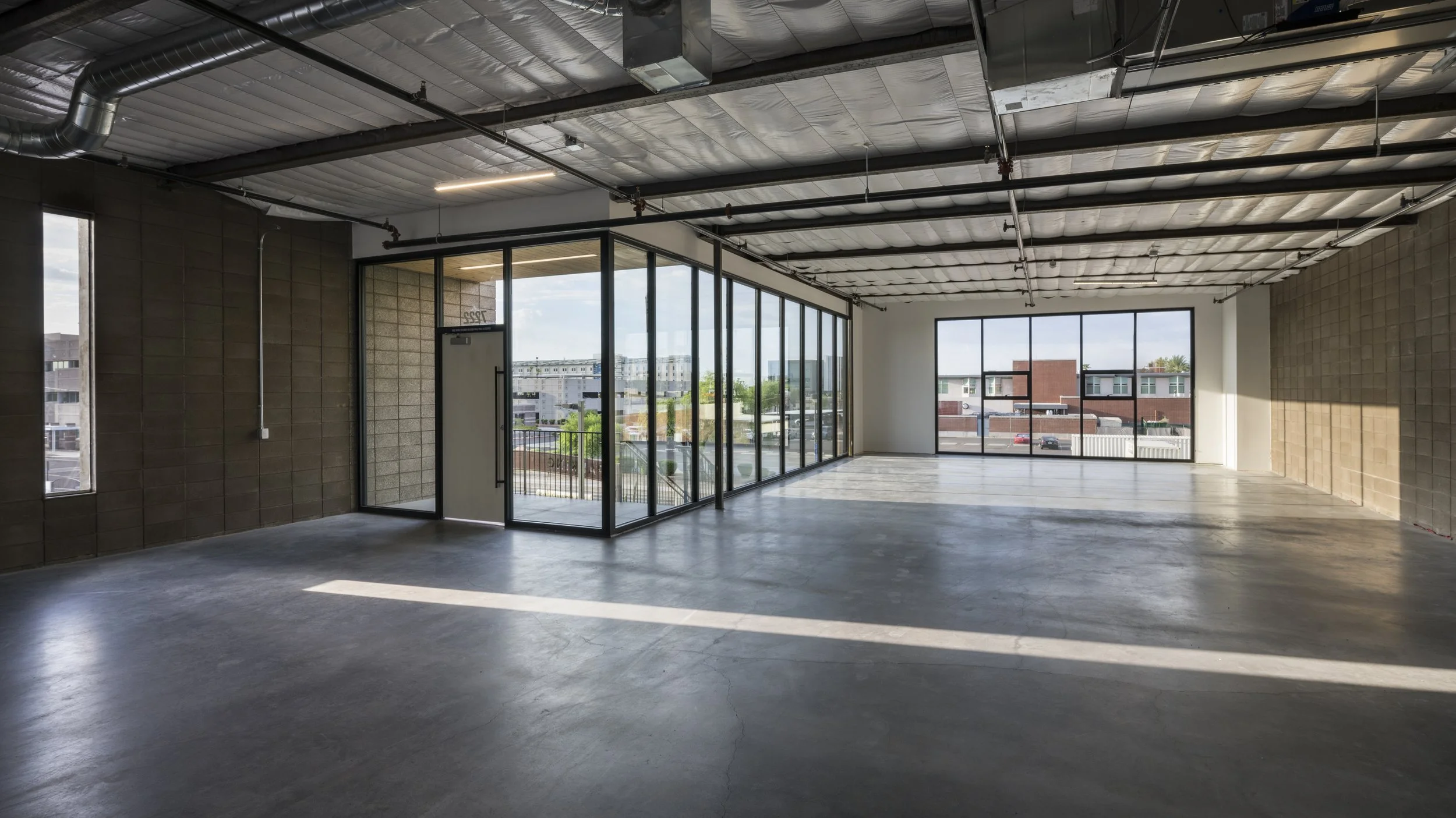 Empty commercial space with large windows, concrete floor, exposed ceiling, and a glass-walled office area.