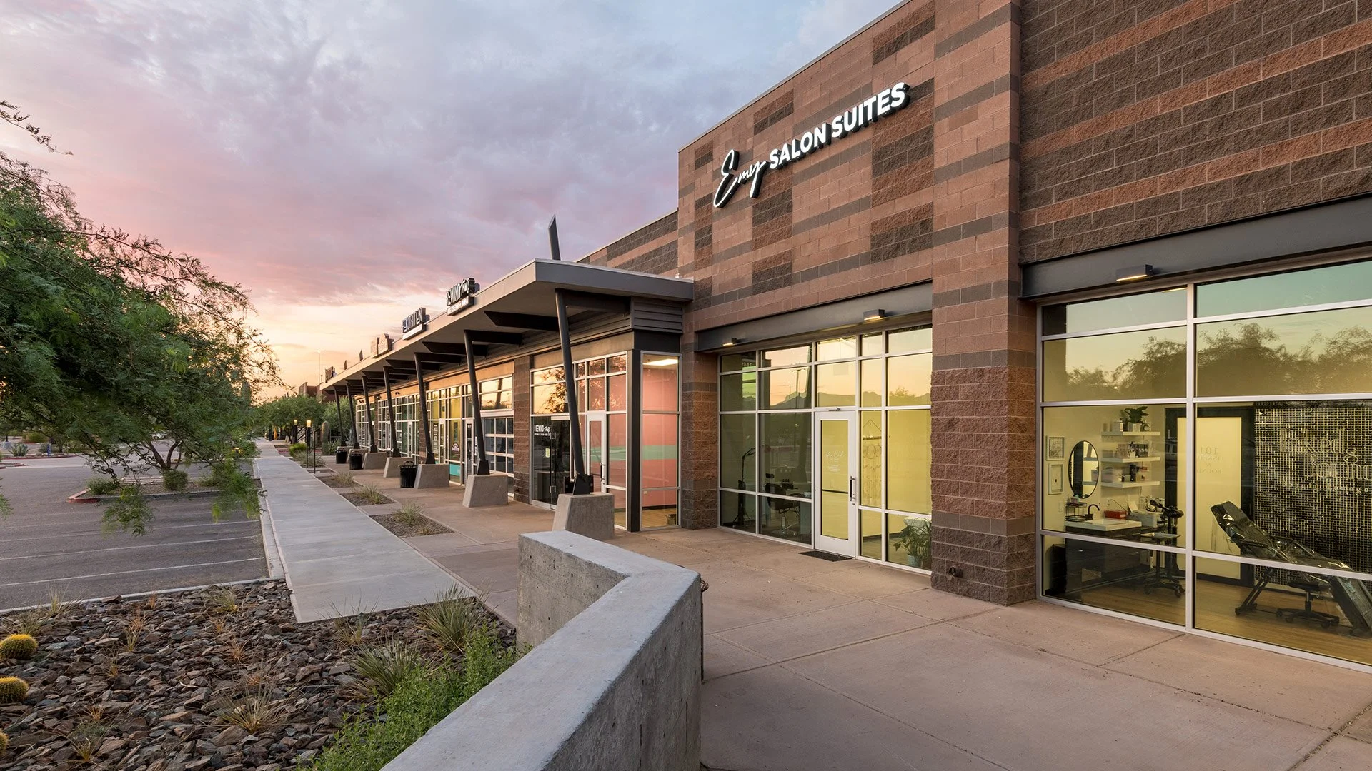 Empty commercial storefront with large glass windows, parking lot, sidewalk, trees, and sunset sky.