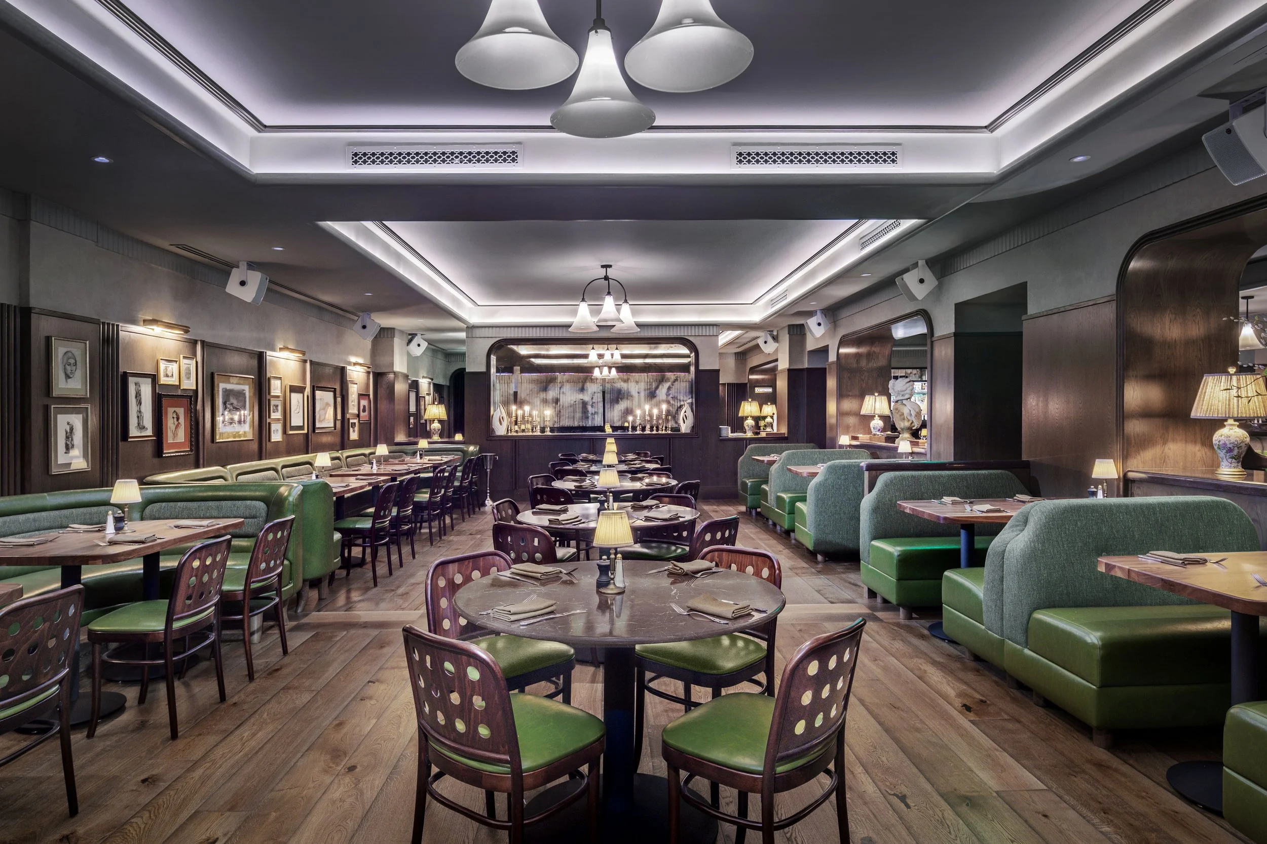 Interior of a restaurant with wooden floors, green booths, and tables set with plates, napkins, and silverware, featuring framed artwork on the walls, small lamps on tables, and ceiling lights.