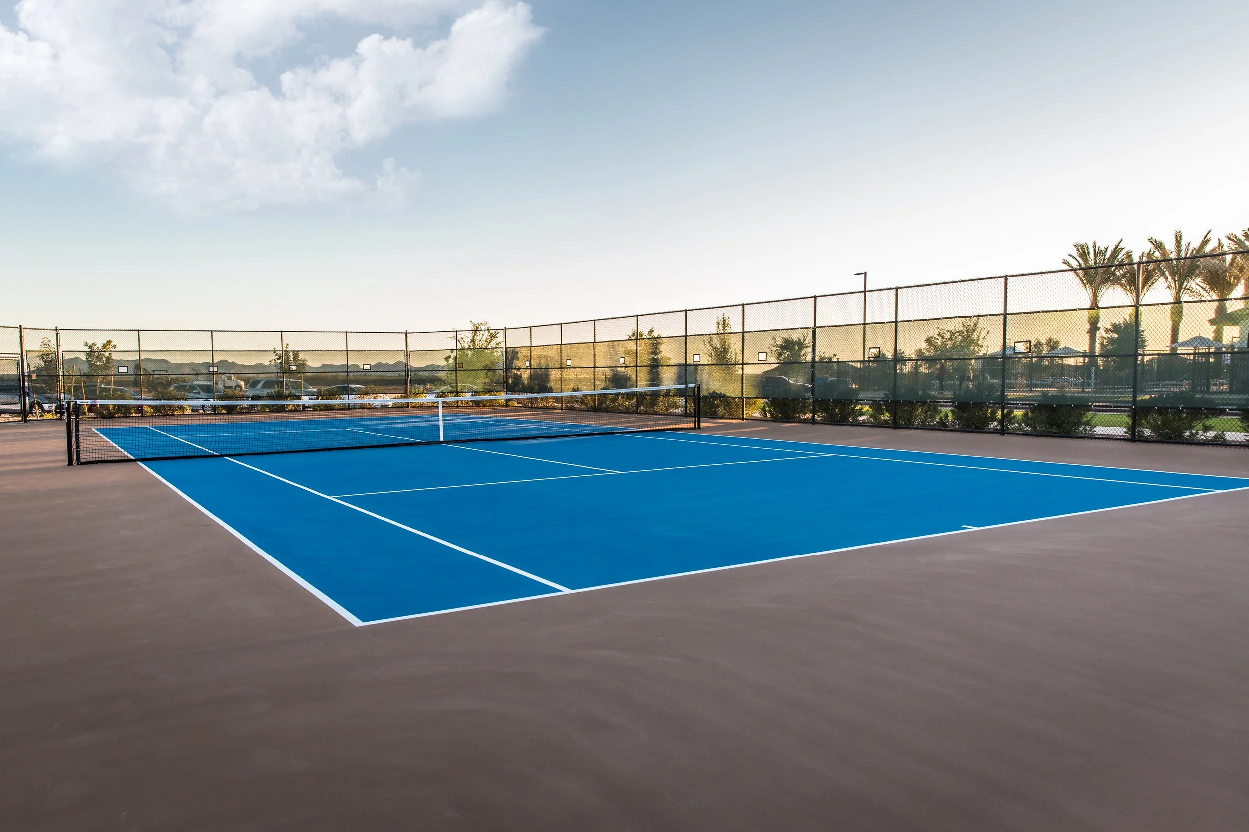Empty blue tennis court with a net, surrounded by a chain-link fence against a sunset sky.
