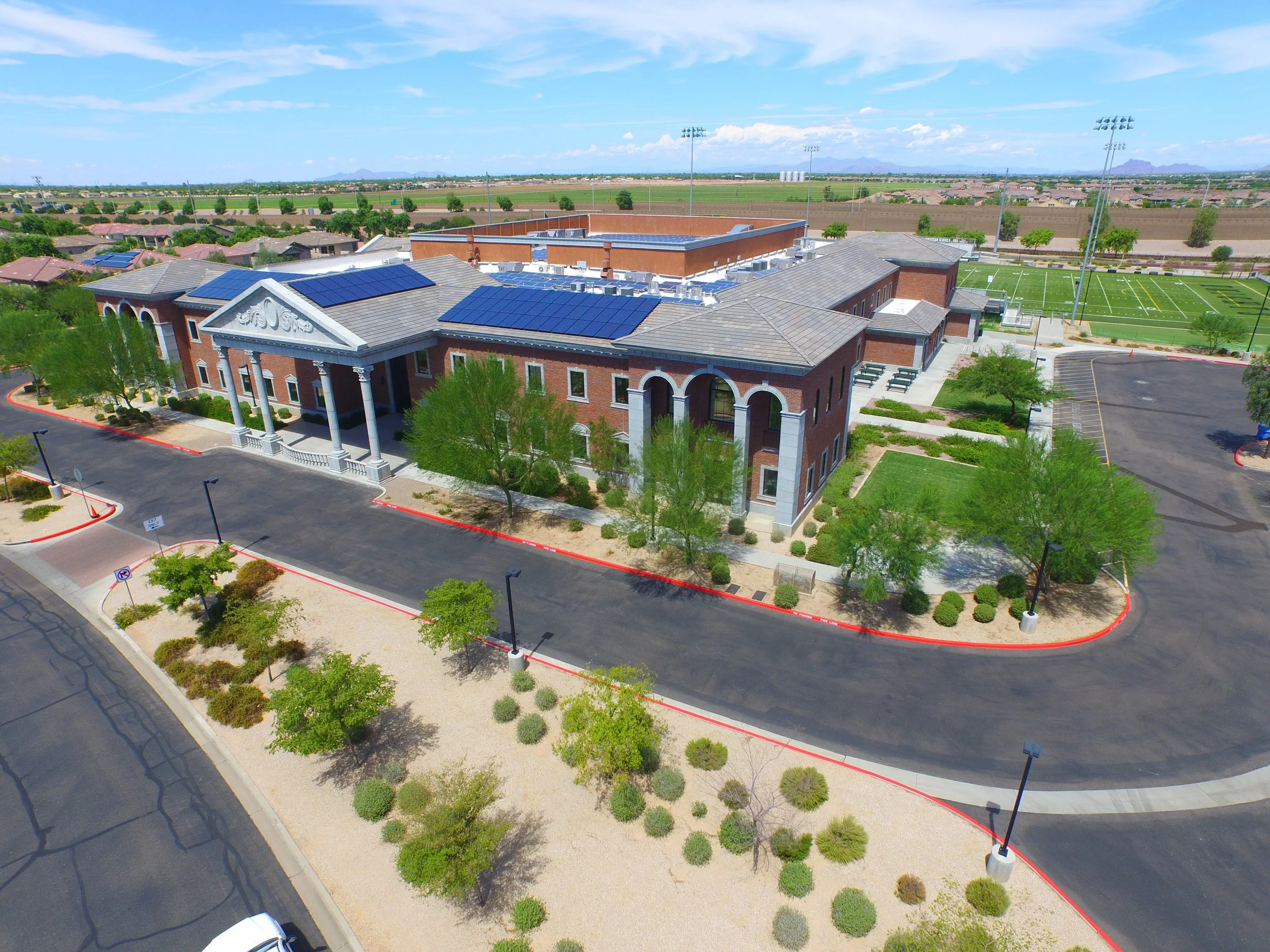Aerial view of a school building with brick façade, solar panels, and surrounding parking lot, green sports field, and desert landscape in the background.