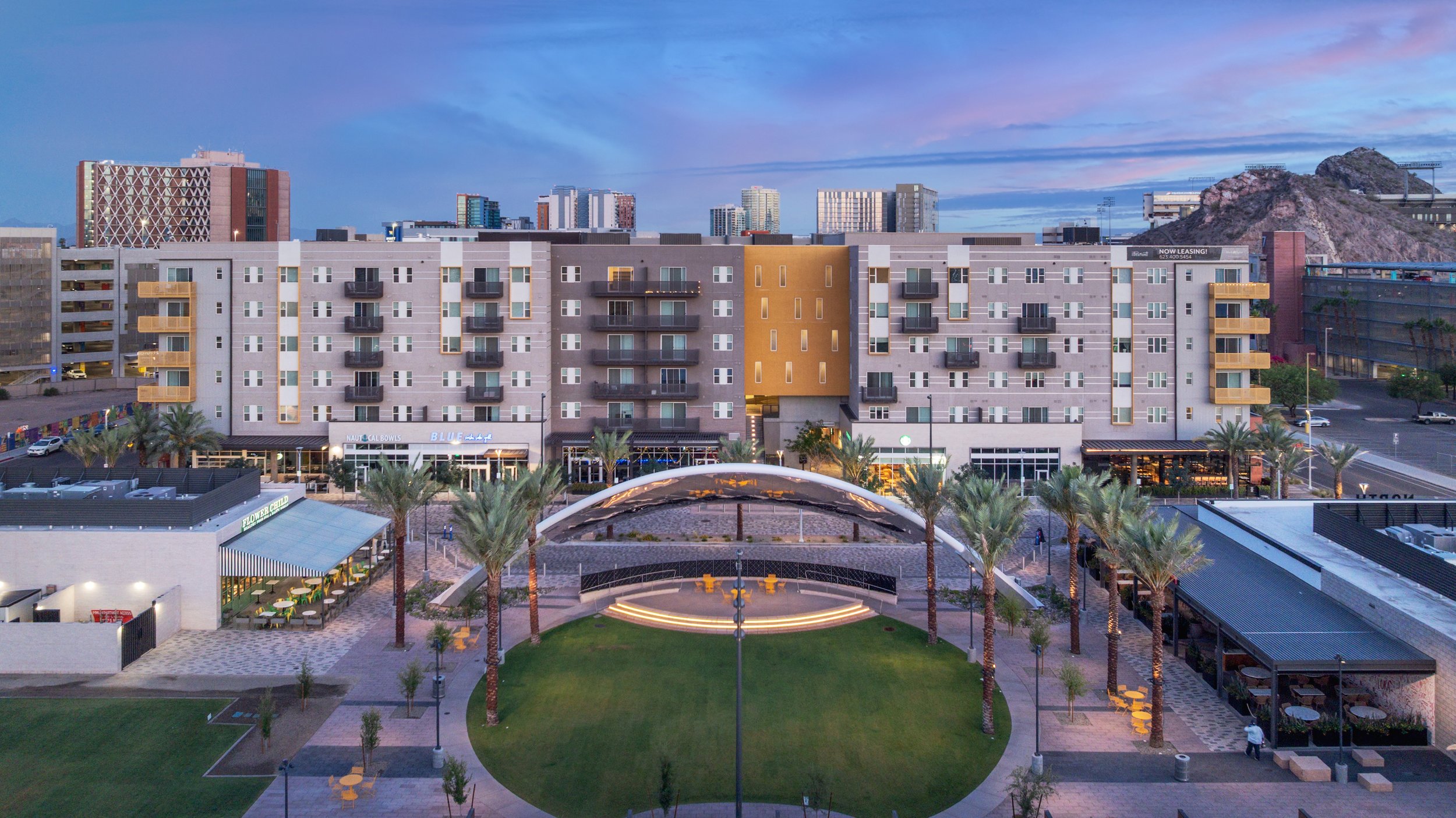 A modern urban plaza with palm trees, a grassy circle, an amphitheater, and surrounding buildings under a twilight sky.
