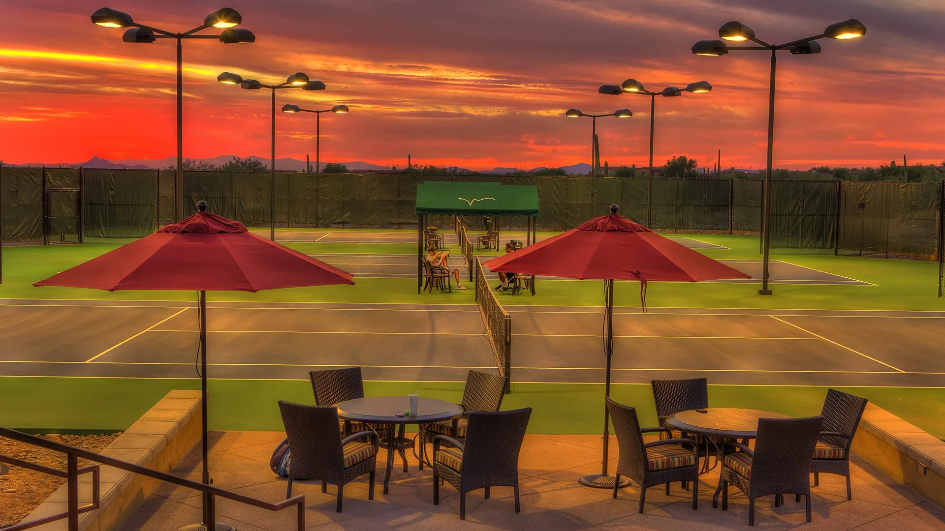 Tennis courts at sunset with outdoor seating and large red umbrellas on a patio.
