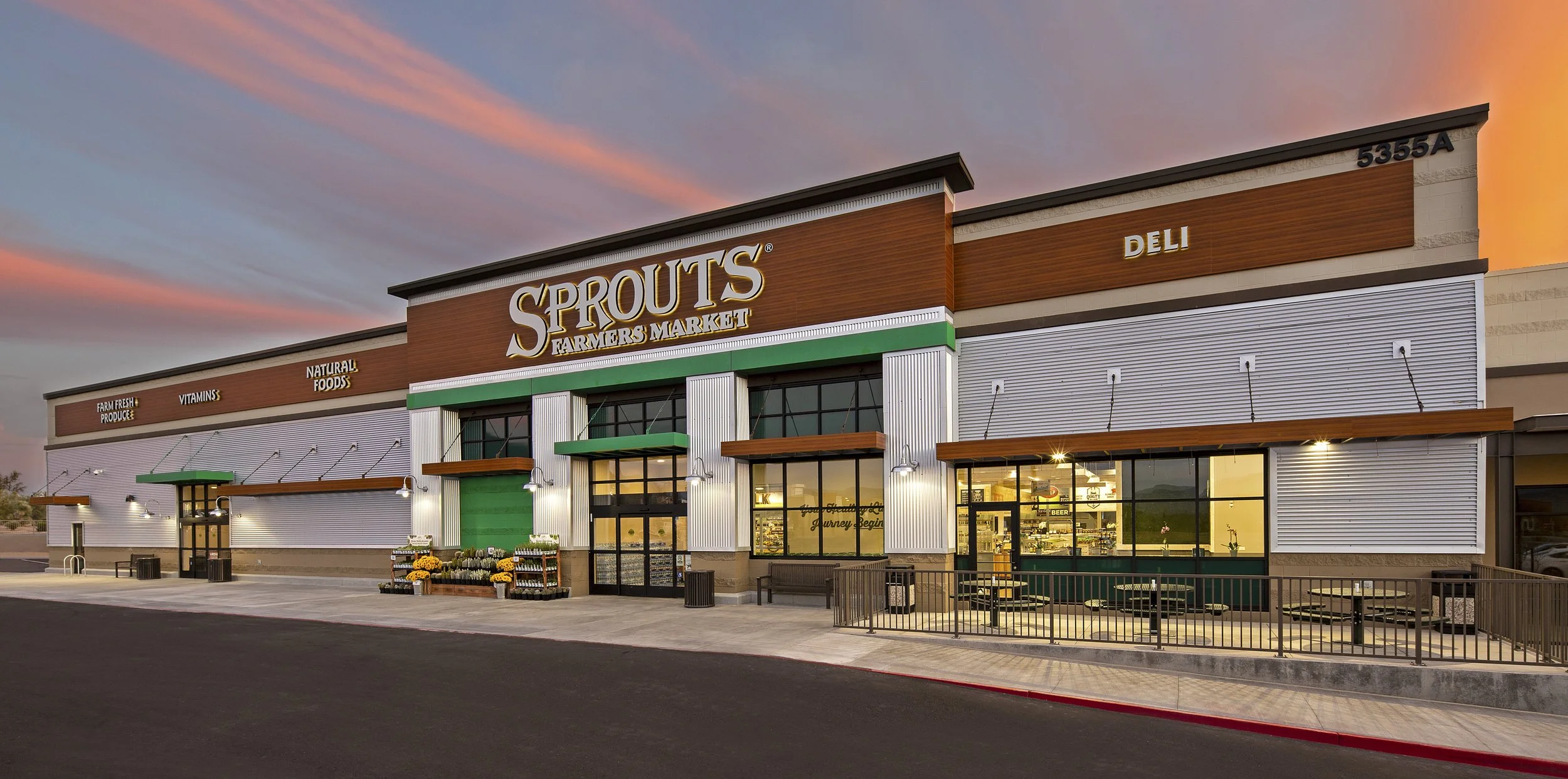 Exterior of Sprouts Farmers Market grocery store at sunset with a colorful sky, large sign, green accents, and outdoor seating area.