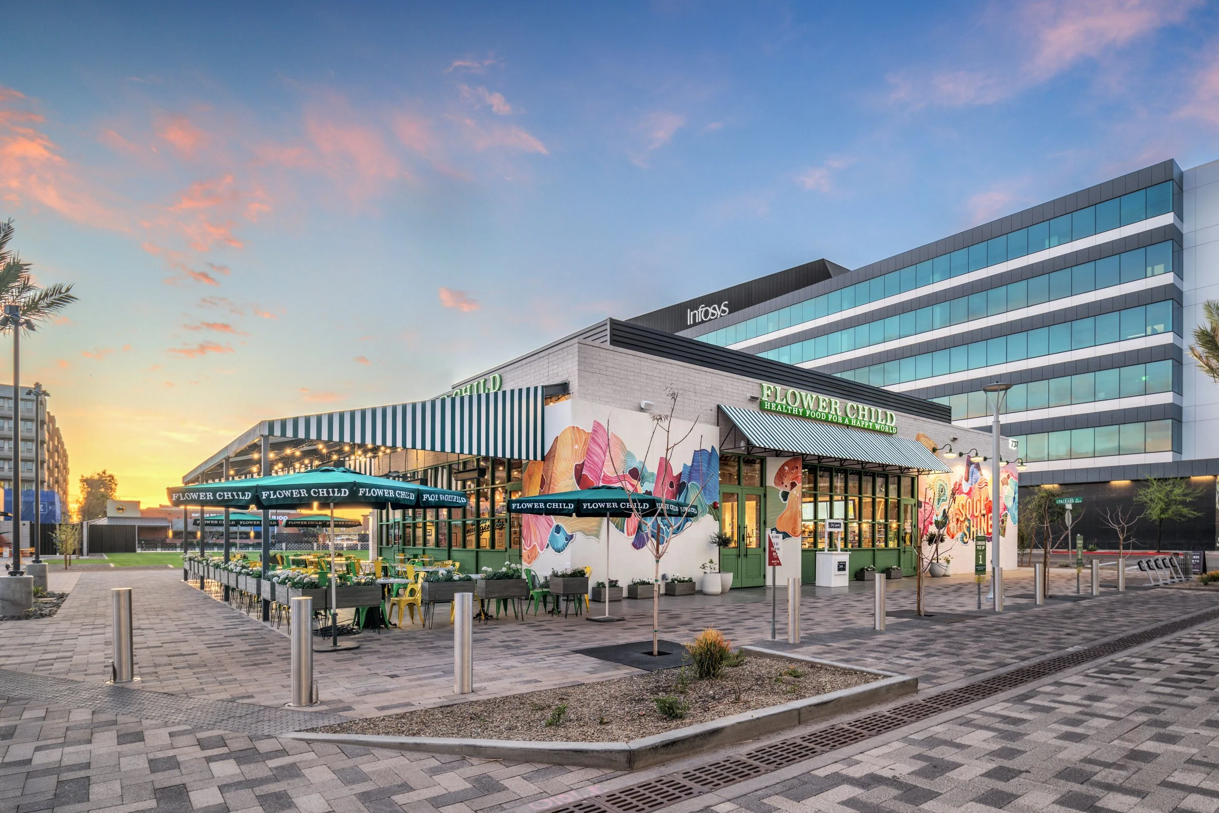 A colorful storefront named 'Flower Child' with outdoor seating, umbrella shades, a mural on the wall, and modern office buildings in the background at sunset.