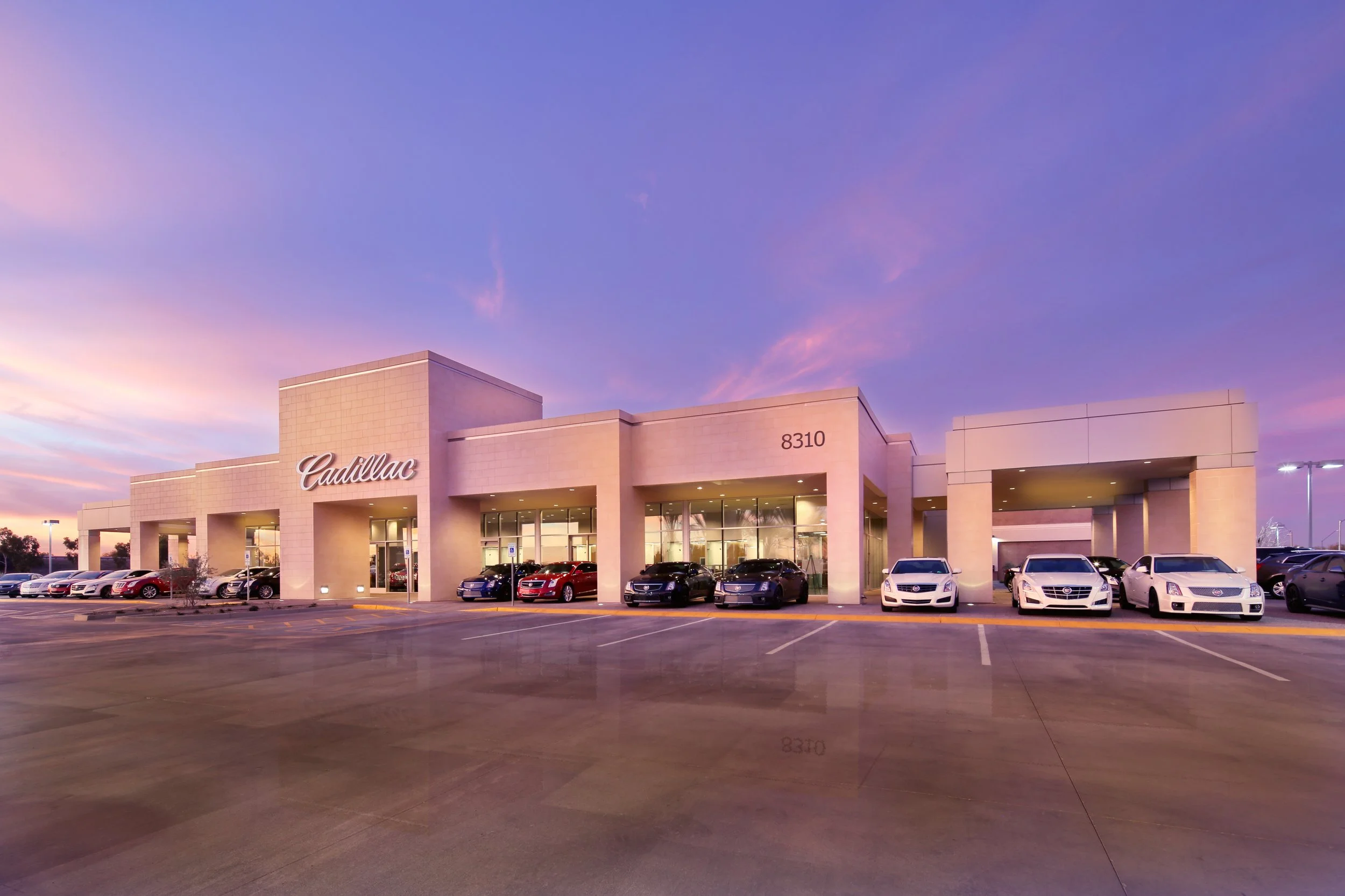 Exterior of Arrowhead Cadillac dealership building with several cars parked in front under a colorful evening sky.